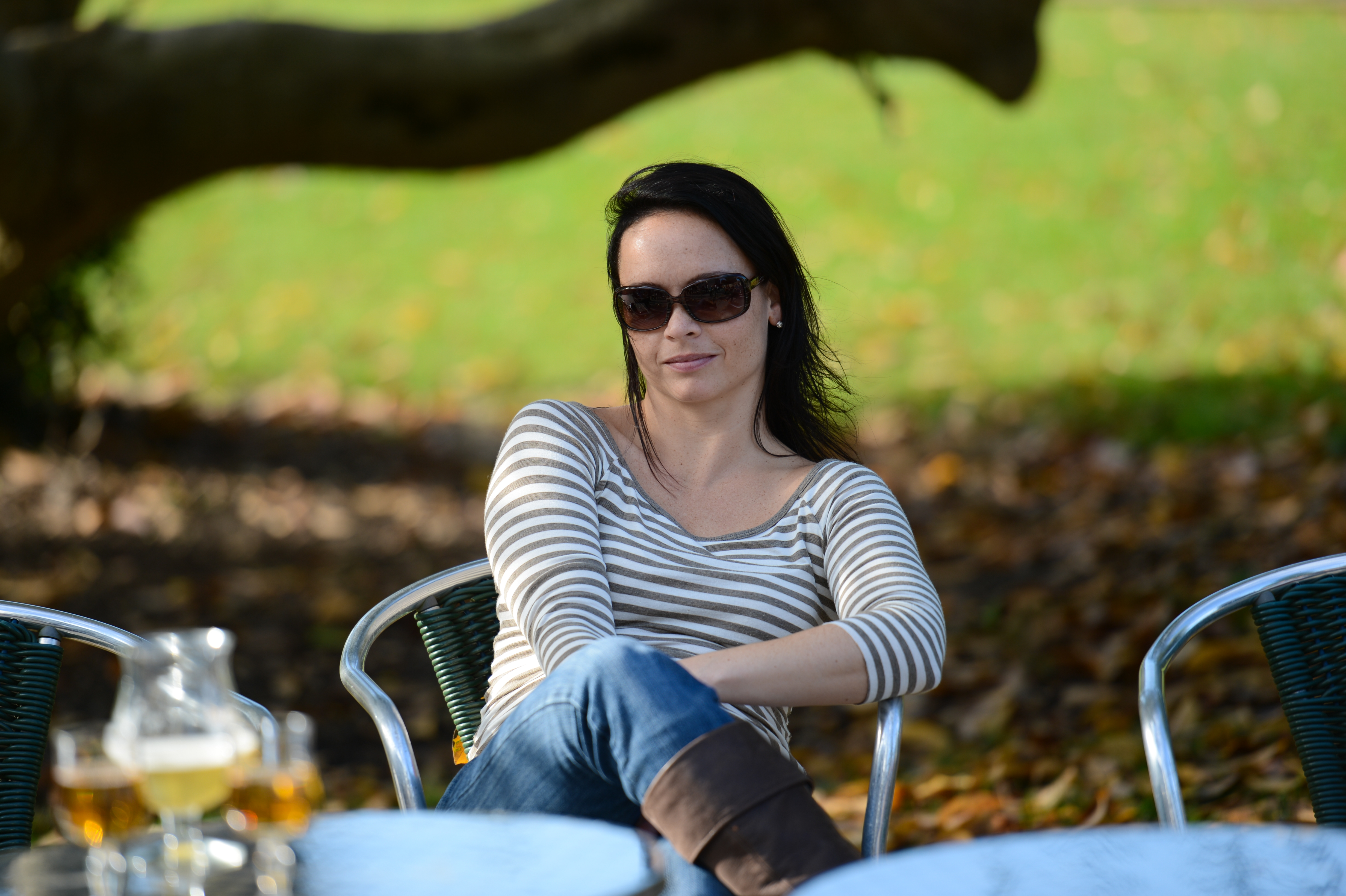 A woman wearing sunglasses sits at an outdoor table, smiling and relaxing in a park-like setting.