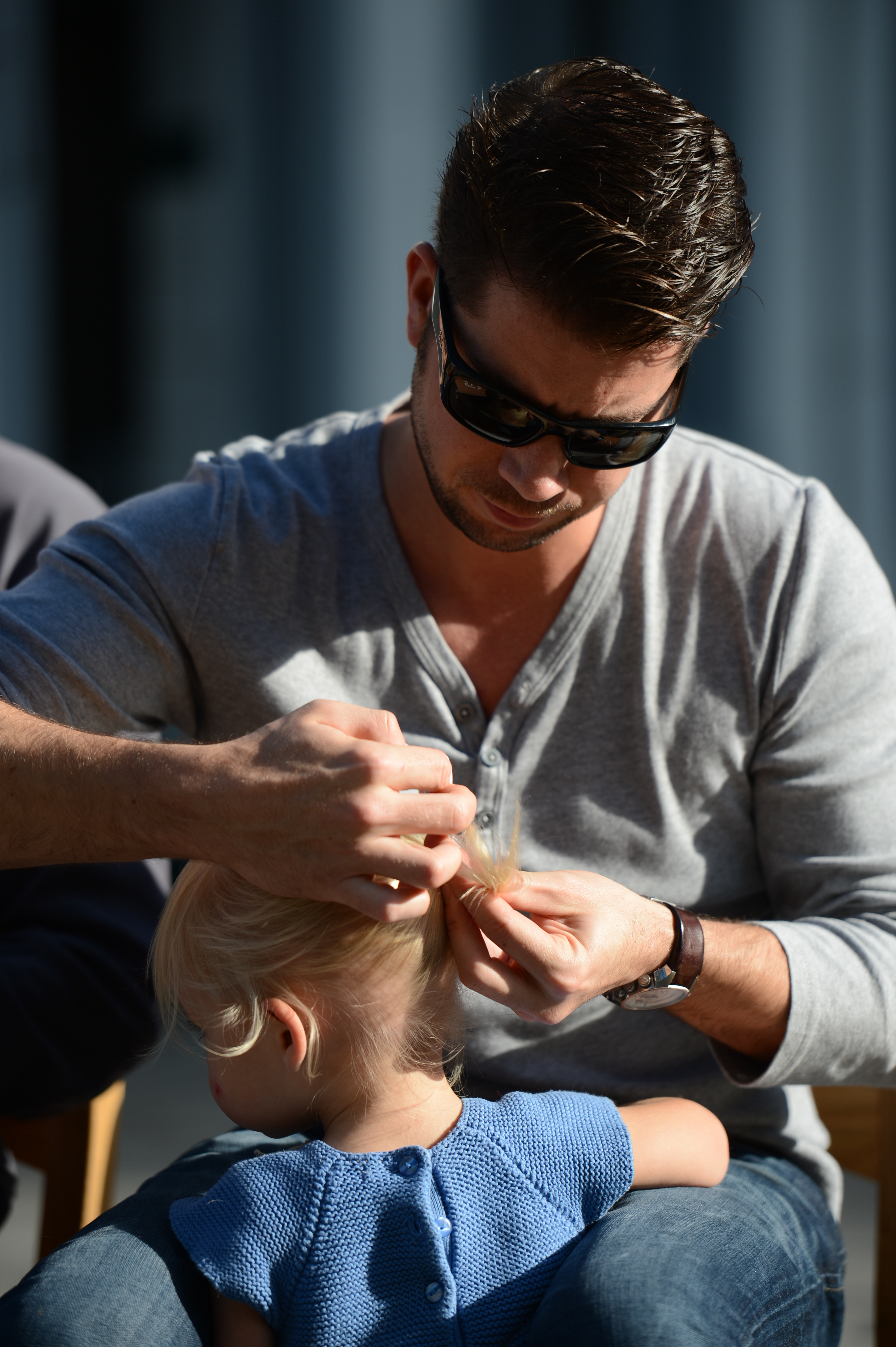 A man wearing sunglasses carefully ties a young child's blonde hair into a small ponytail.