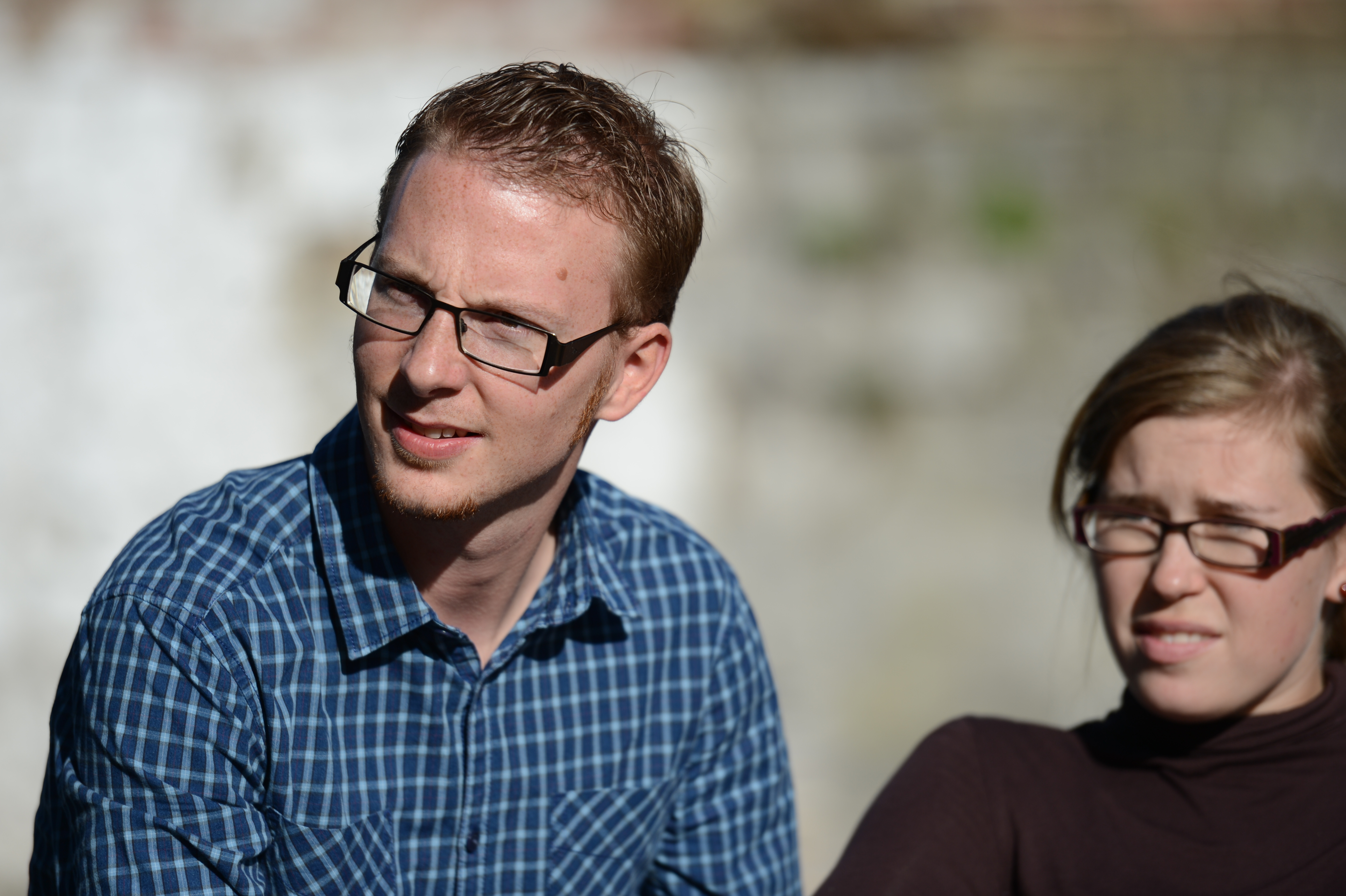 A man and a woman wearing glasses sit outside, squinting in the sunlight and looking in the same direction.