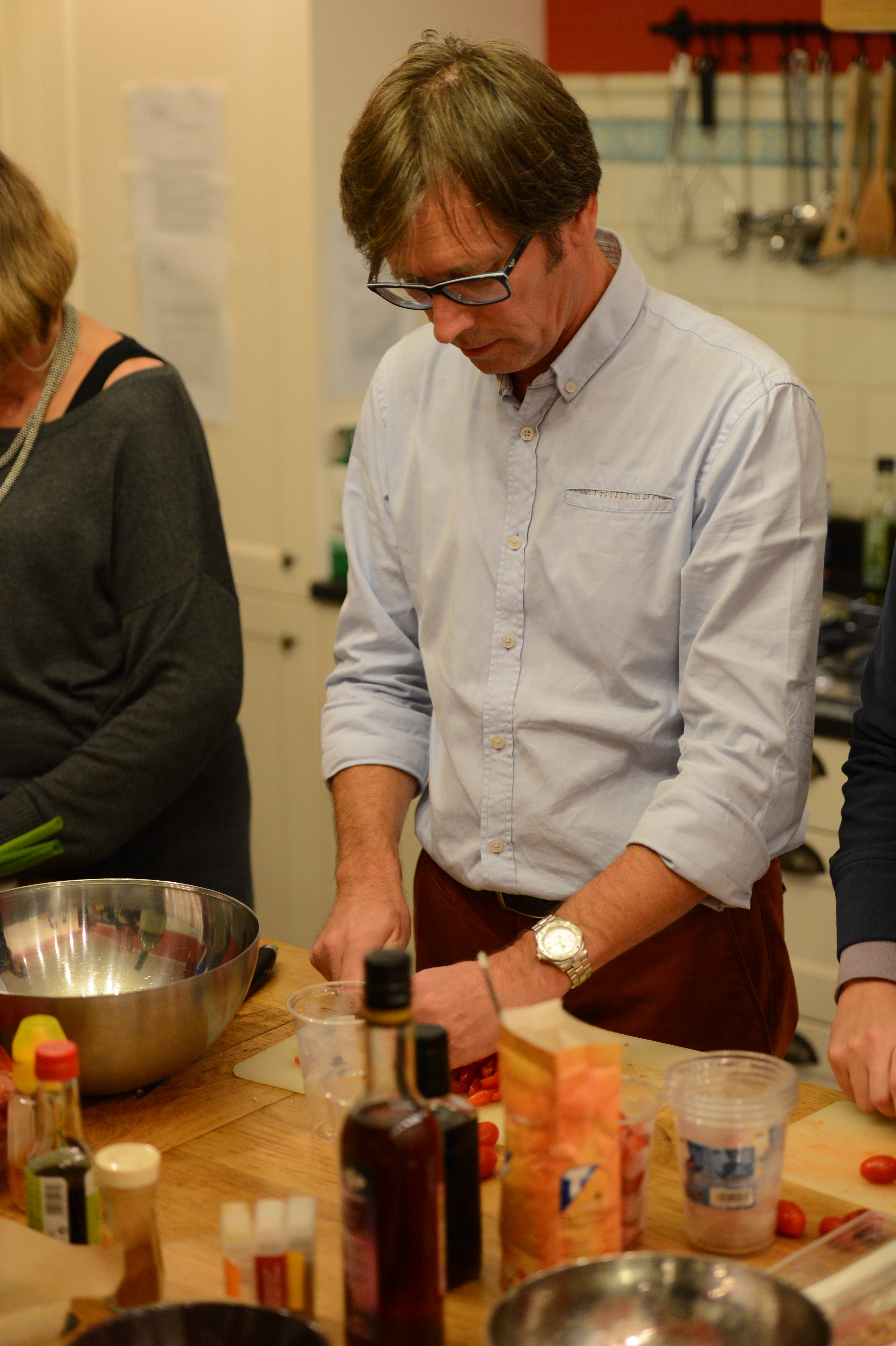 A man in glasses and a light blue shirt chops food on a cutting board during a family cooking activity.