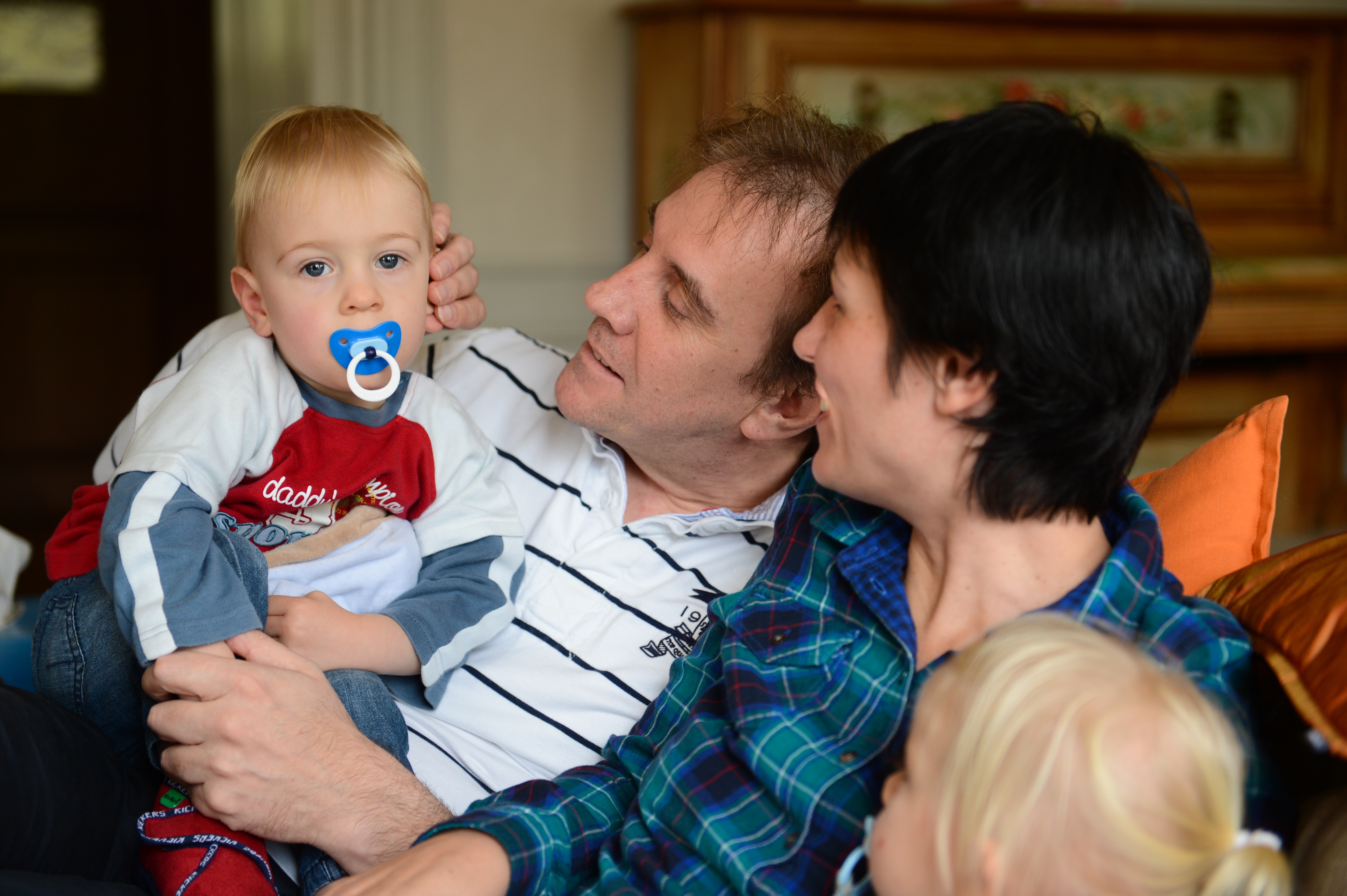 A family sits together on a couch, with parents smiling and holding their young children.