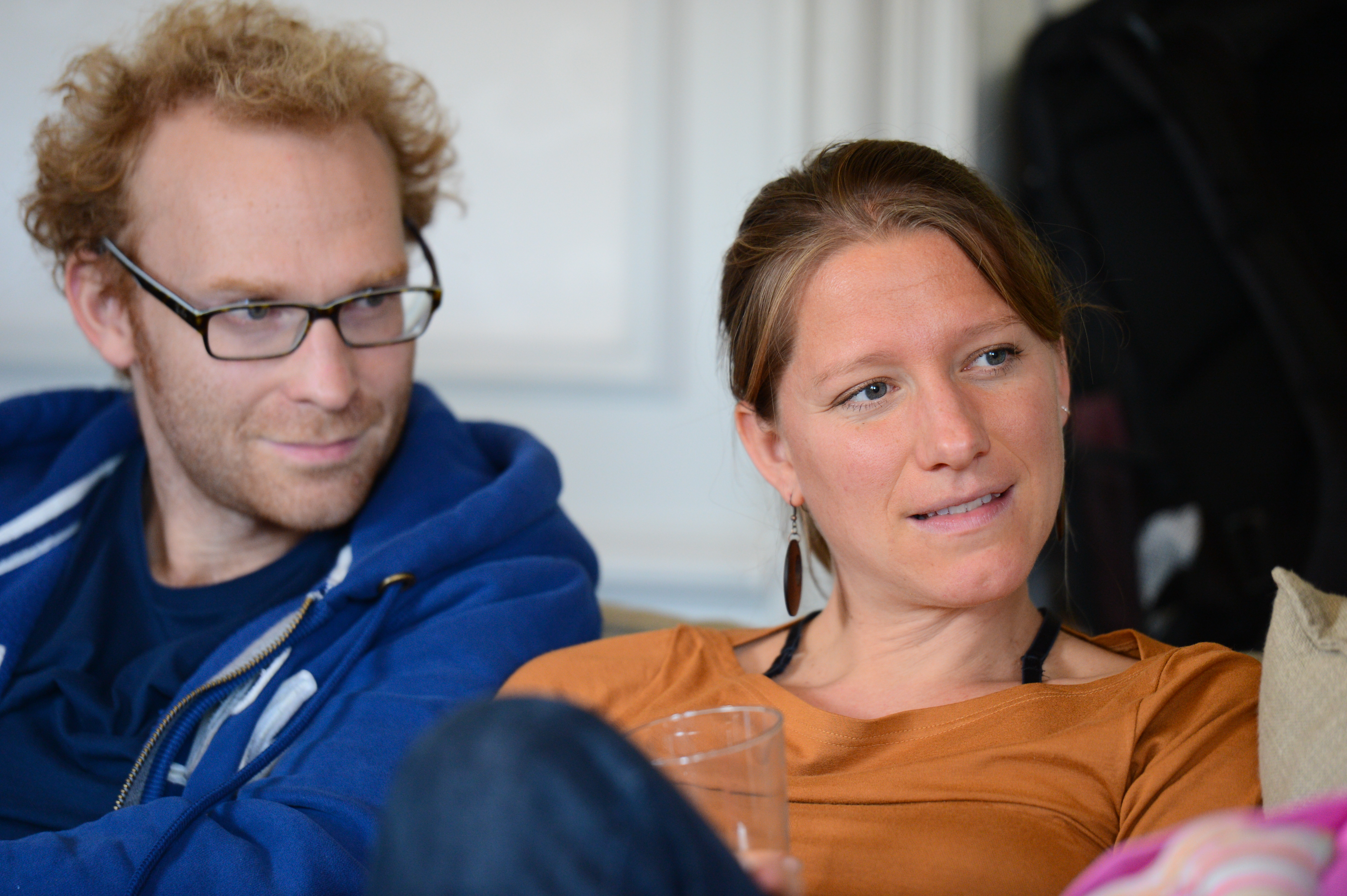 A man in a blue hoodie and a woman in a brown top sit together, engaged in conversation.