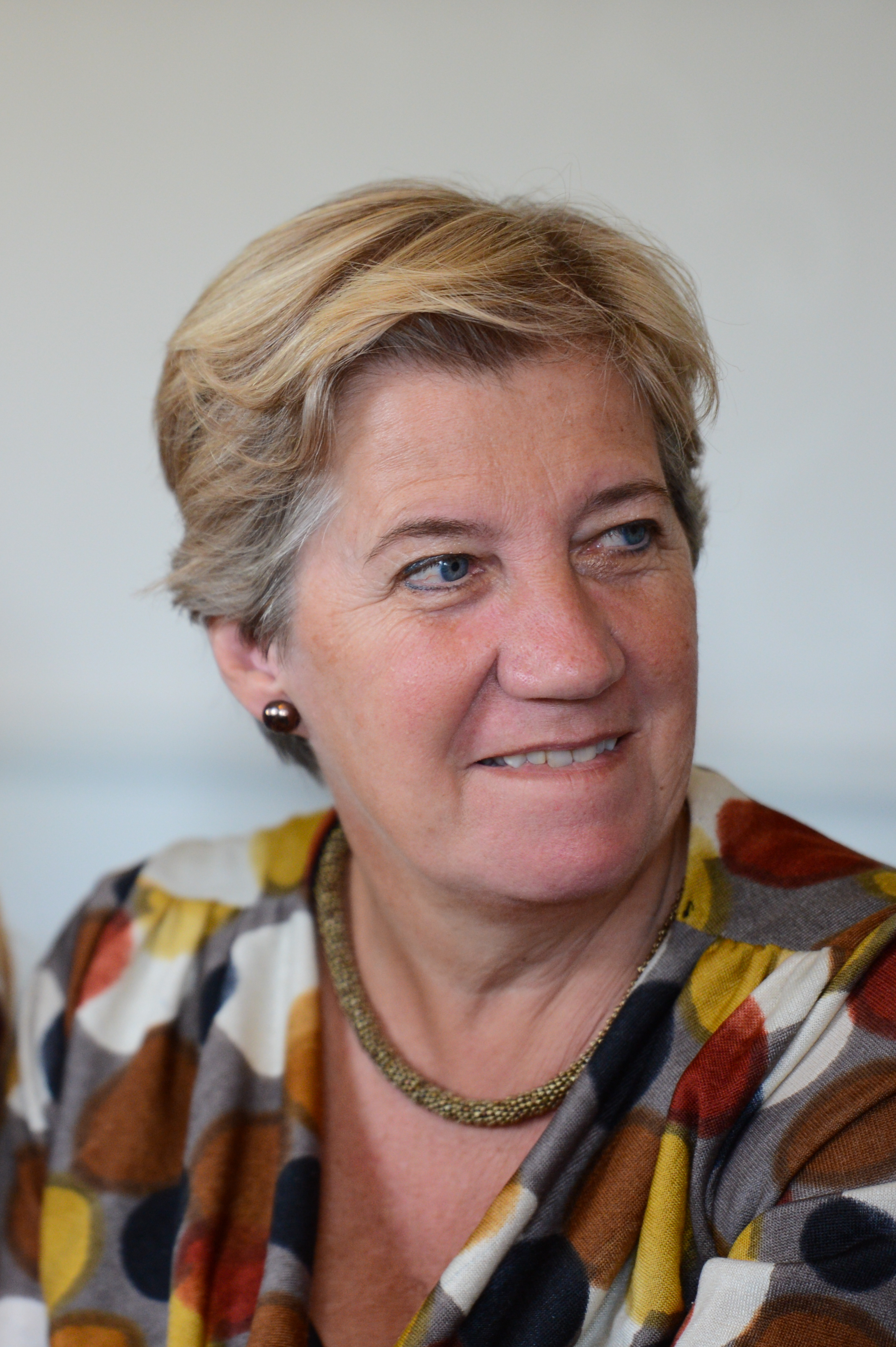 A woman with short blonde hair and a patterned blouse looks to the side with a slight smile.