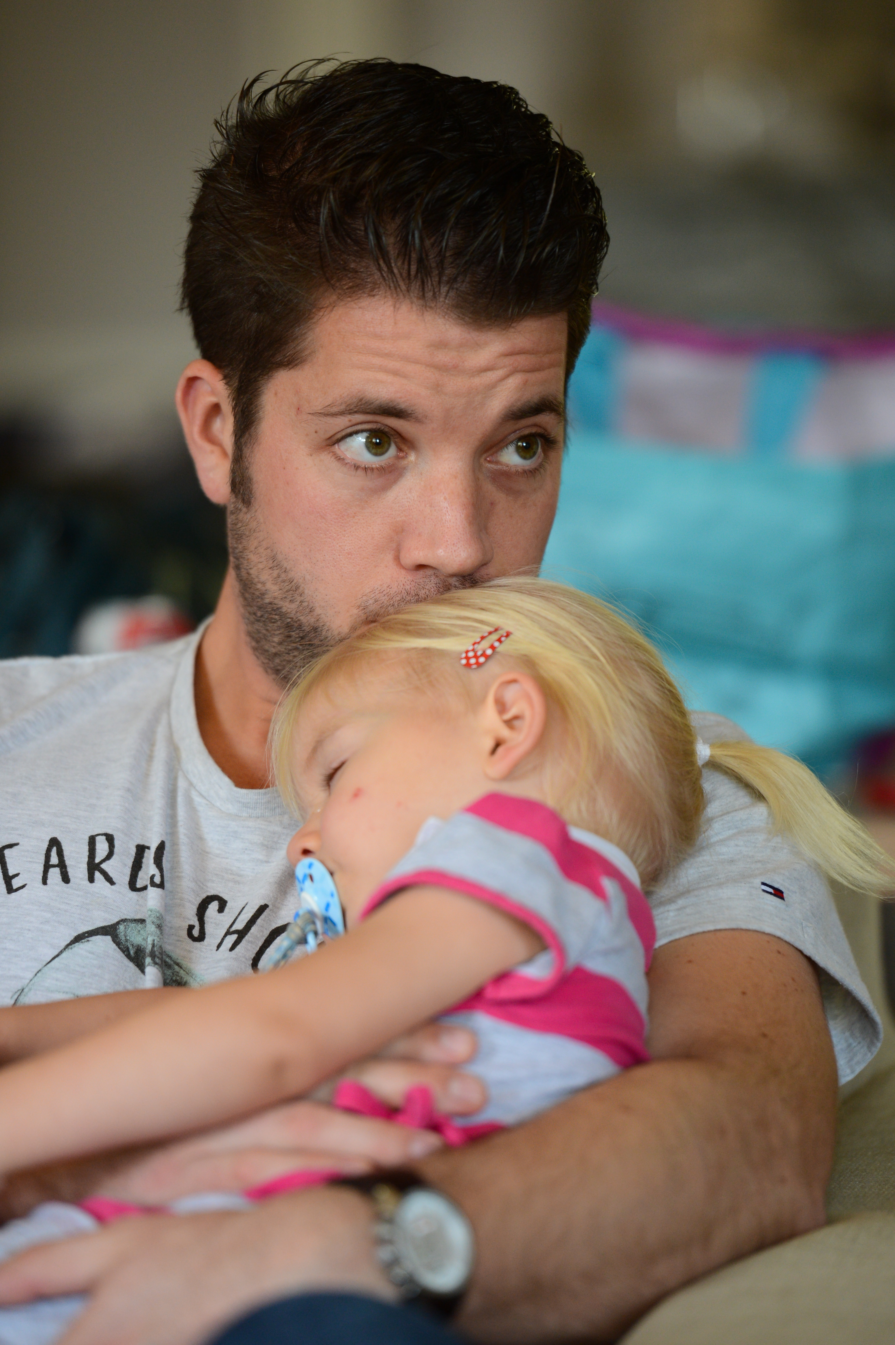 A man sits on a couch, holding a sleeping toddler in his arms while looking into the distance.