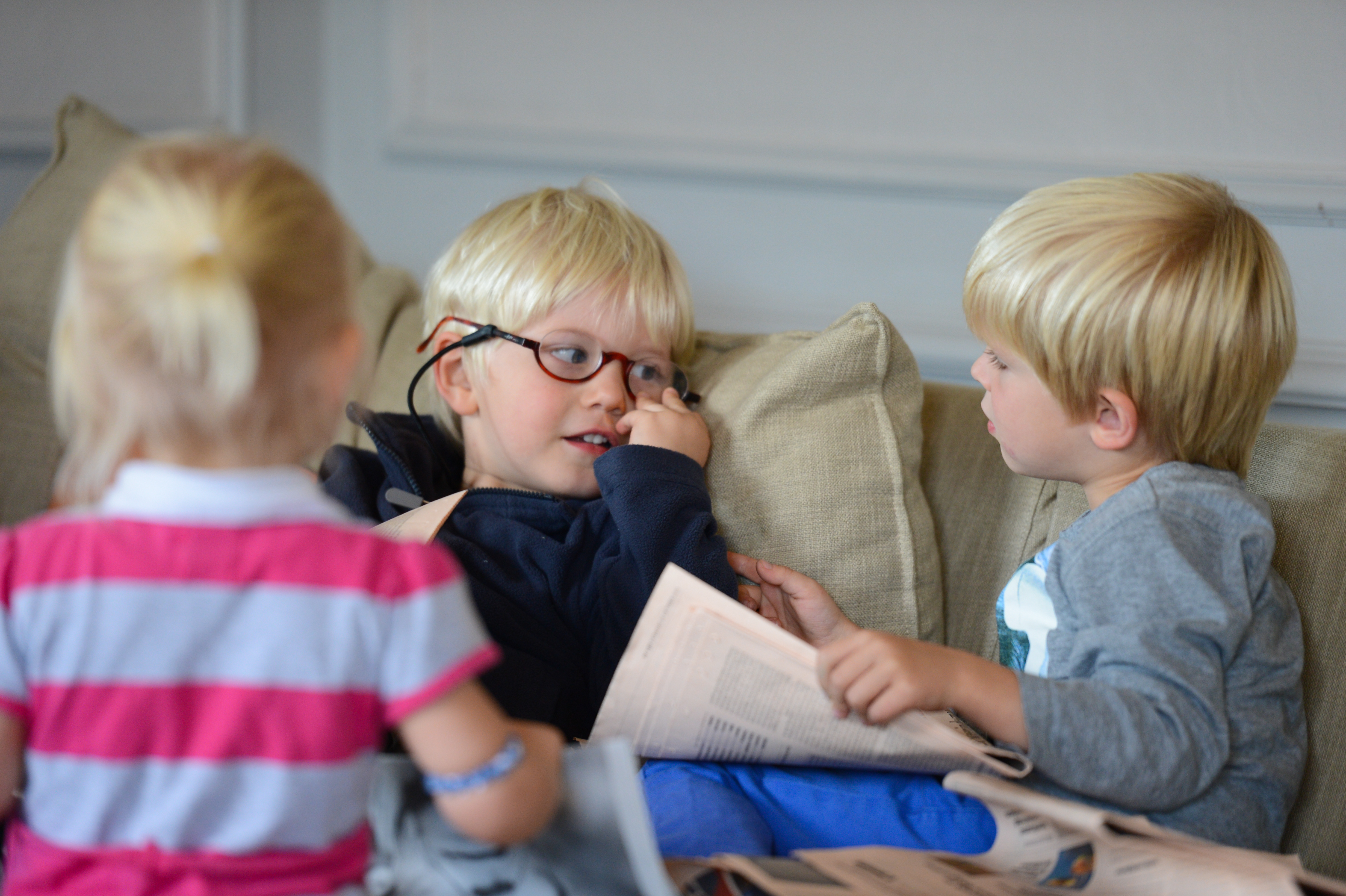 Three young children with blonde hair sit on a couch, talking and playing with a newspaper.