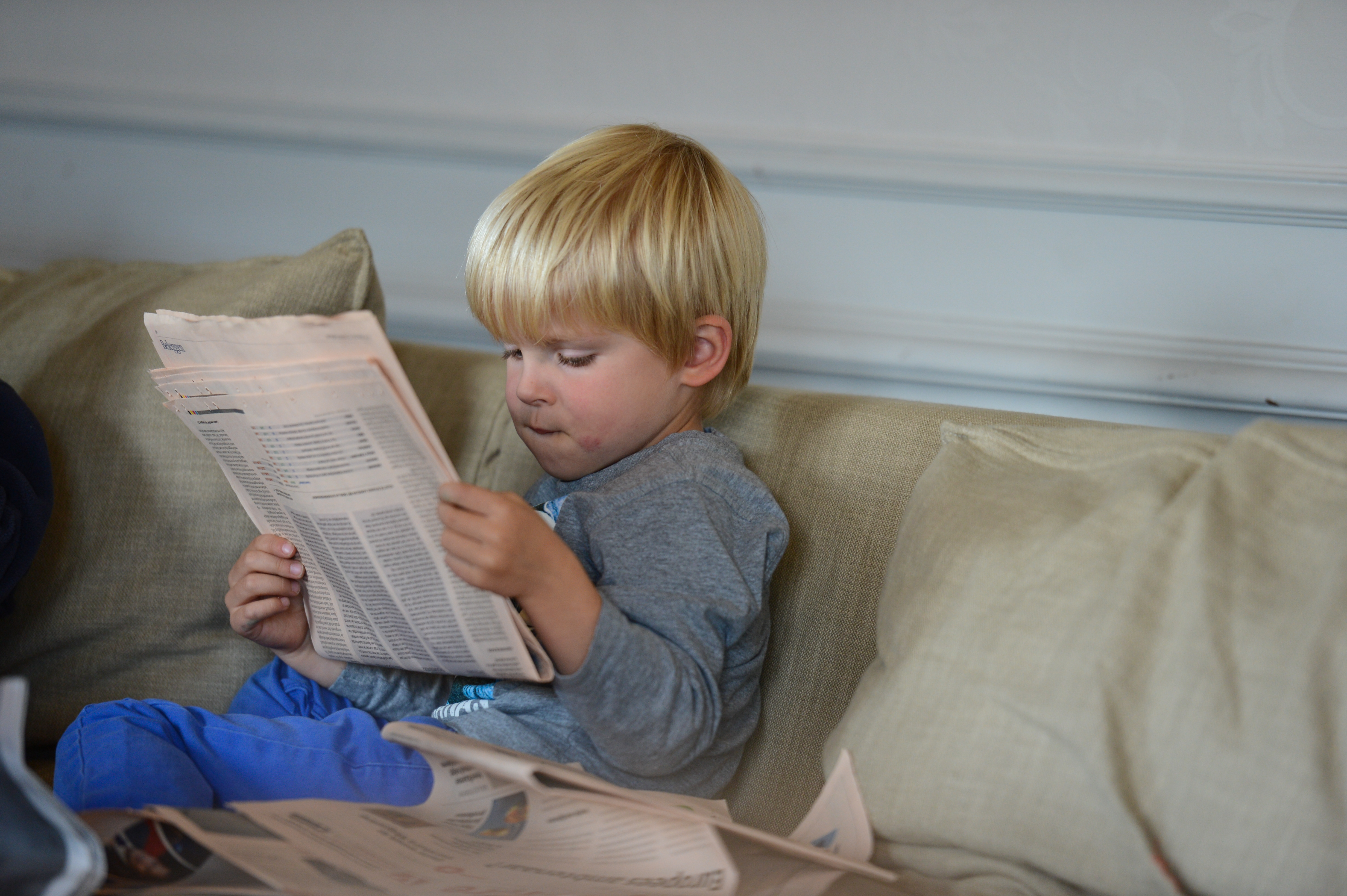 A young child sits on a couch, focused on reading a newspaper.
