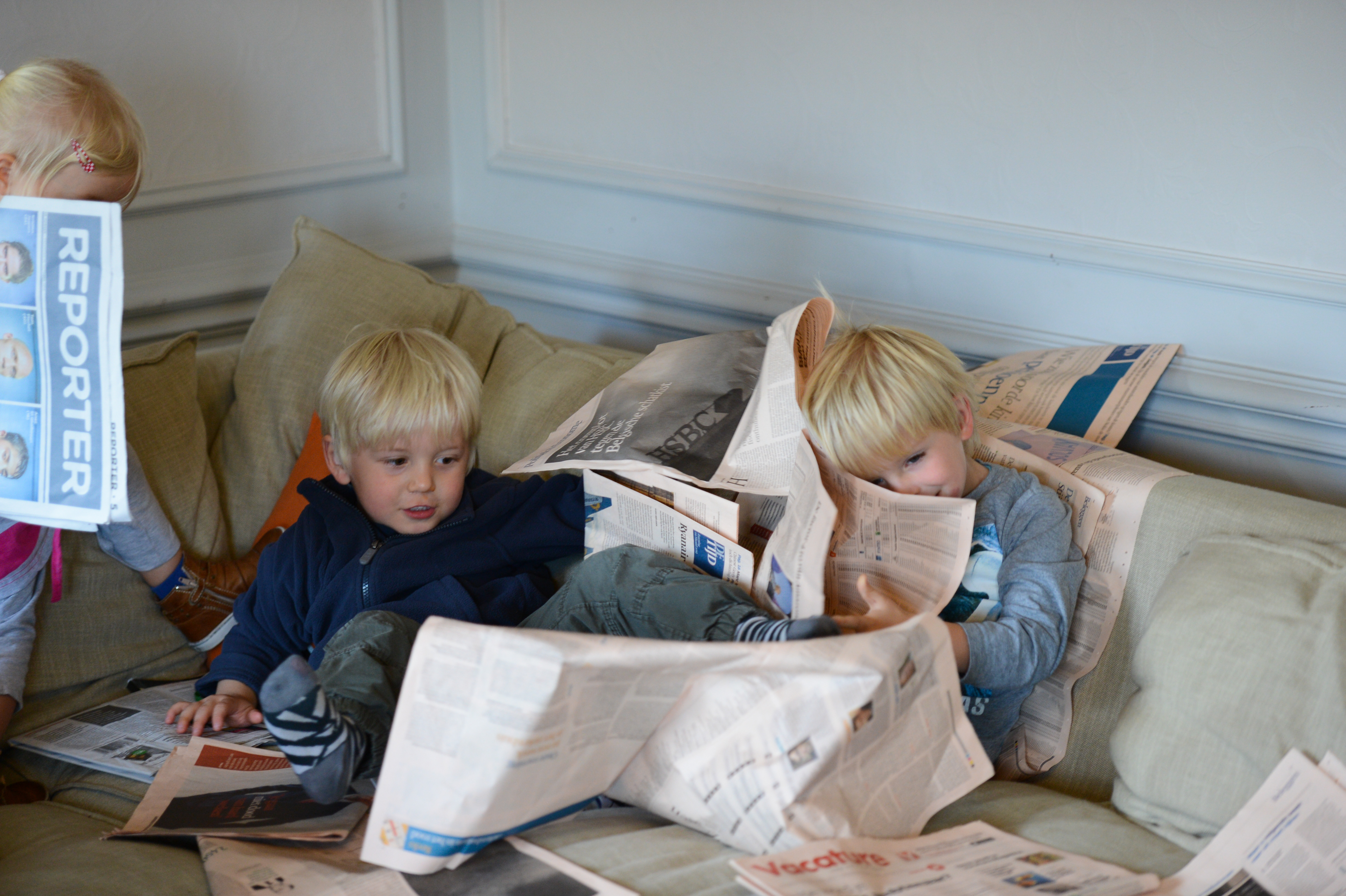 Two young children sit on a couch, playing with newspapers, while another child holds up a newspaper nearby.