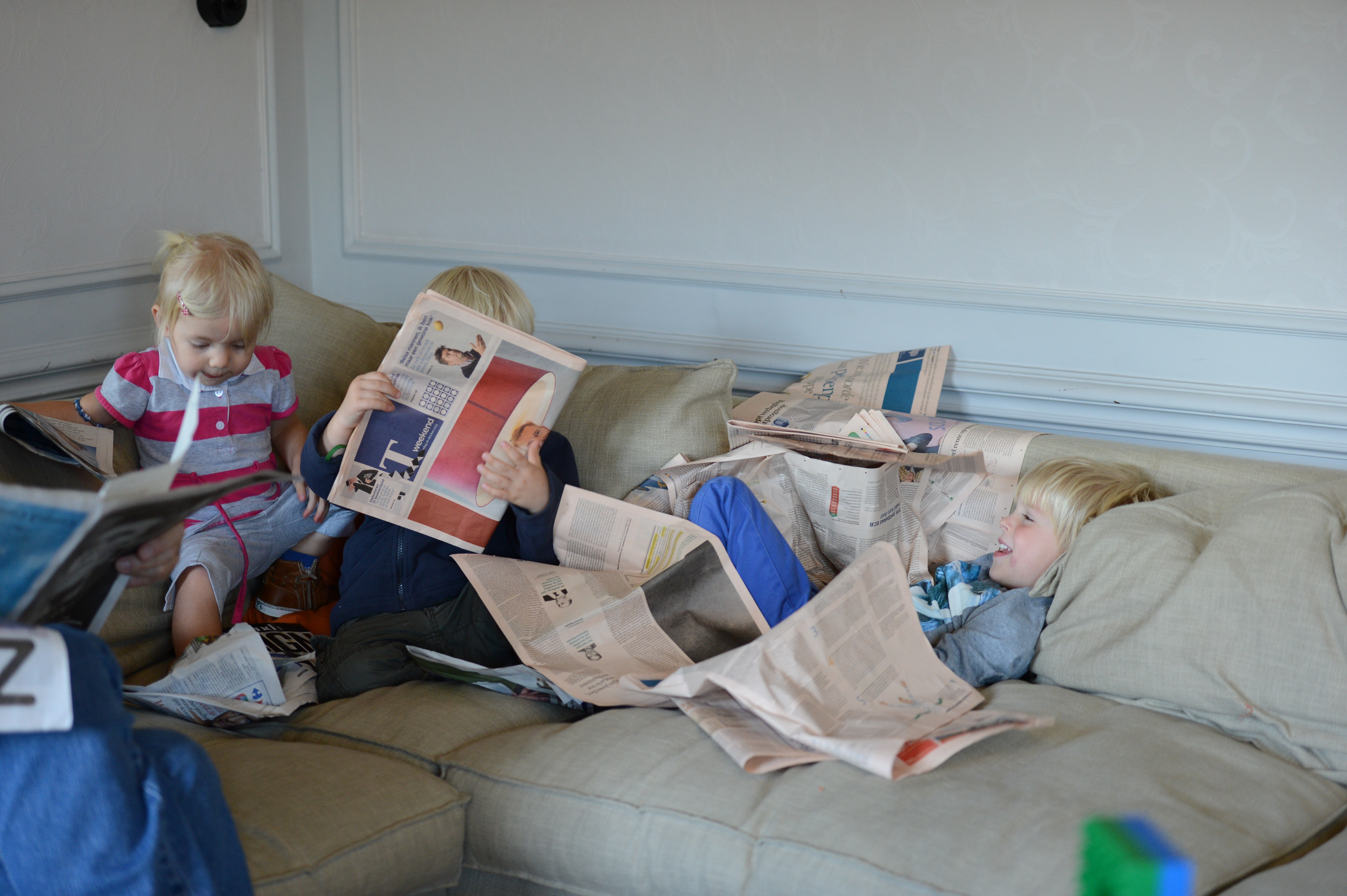 Three young children sit on a couch, playing with newspapers and laughing together during a family weekend.