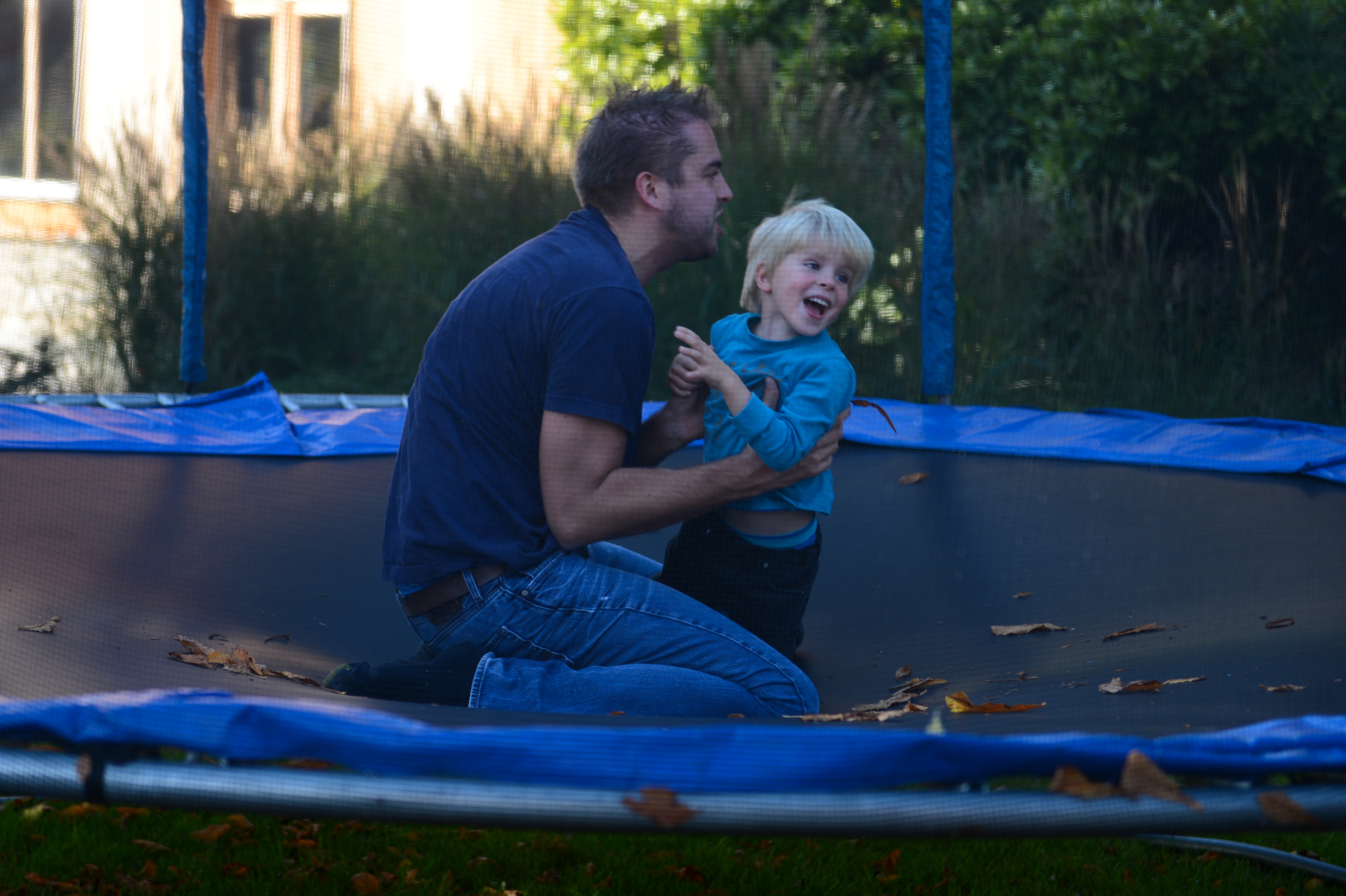A man and a young child laugh together while kneeling on a trampoline outdoors.
