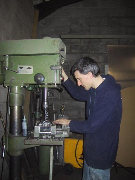 A person operates a large drill press in a workshop, adjusting the machine while focusing on the task.