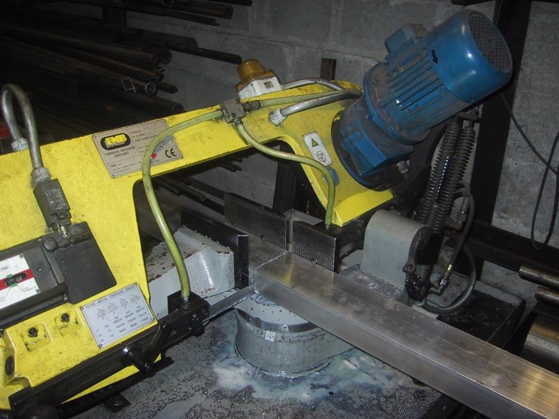 A metal-cutting bandsaw with a yellow frame and blue motor cuts an aluminum bar in an industrial workshop.