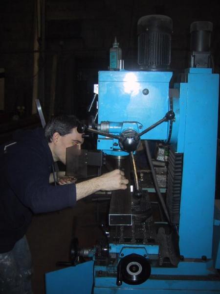 A person operates a blue milling machine, carefully adjusting a metal piece under the drill.