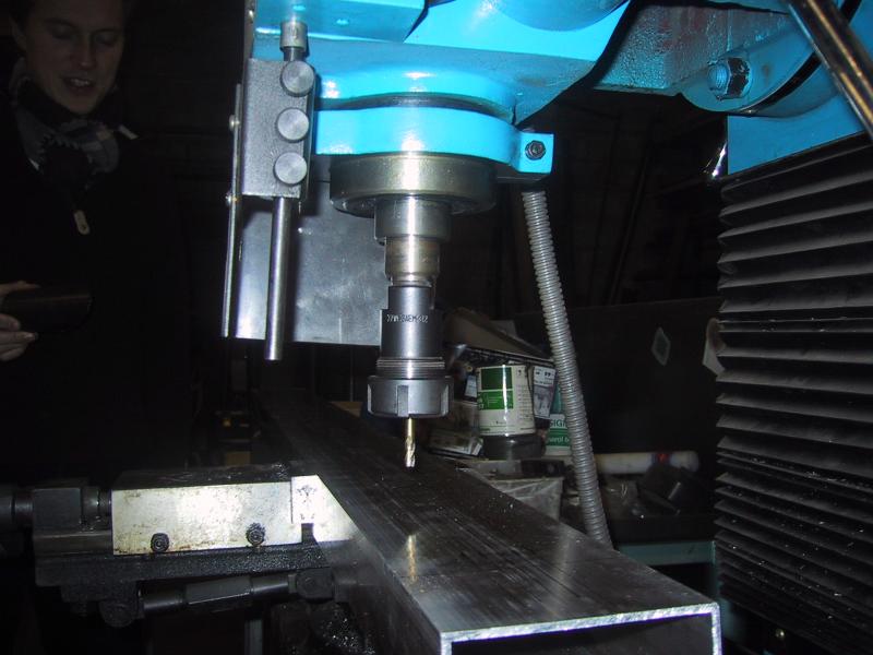 A milling machine drills into a metal beam in a workshop, with a person observing in the background.