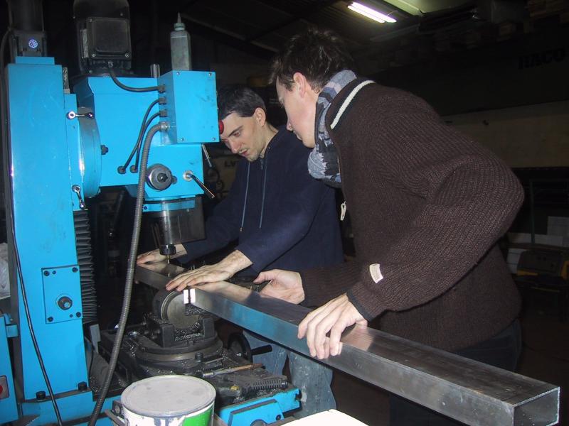 Two people operate a blue industrial machine, guiding a long metal piece into position in a workshop.