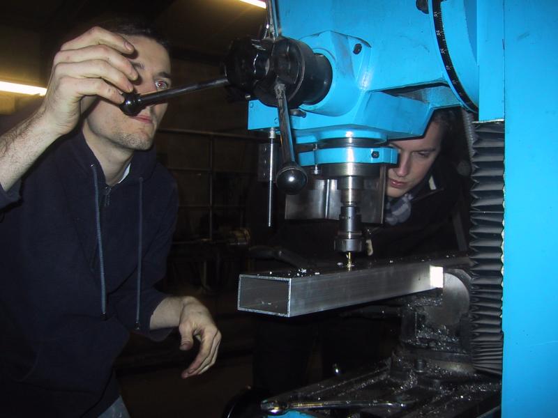 A person operates a blue milling machine while another observes closely, focusing on machining a metal piece.