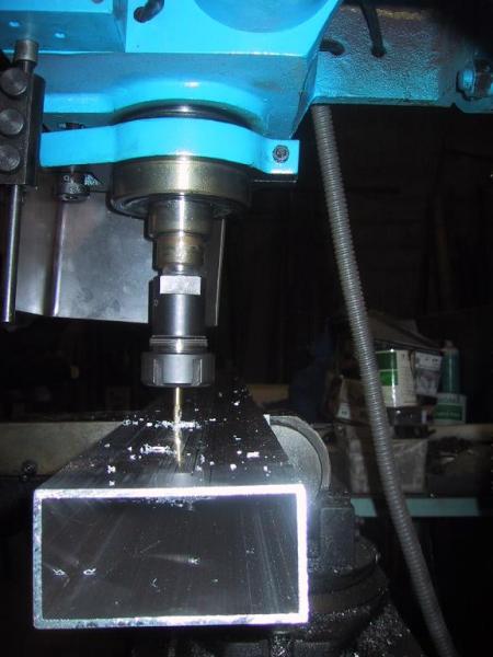 A milling machine drills a hole into a rectangular metal tube, with metal shavings scattered around.