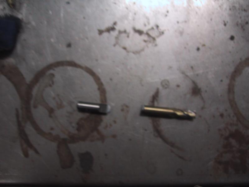 Two small metal drill bits lying on a scratched and stained metallic surface.