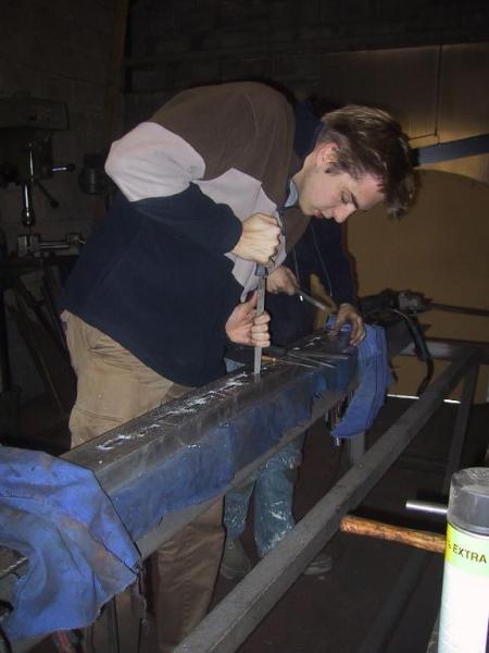 A person uses a file to shape a metal beam in a workshop, with another person working nearby.