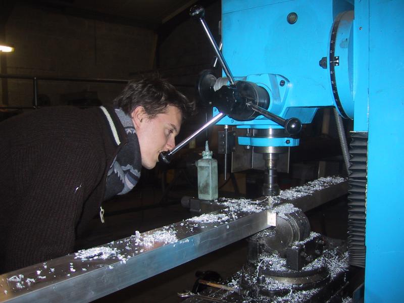 A person closely inspects a milling machine cutting a metal beam, with metal shavings scattered around.
