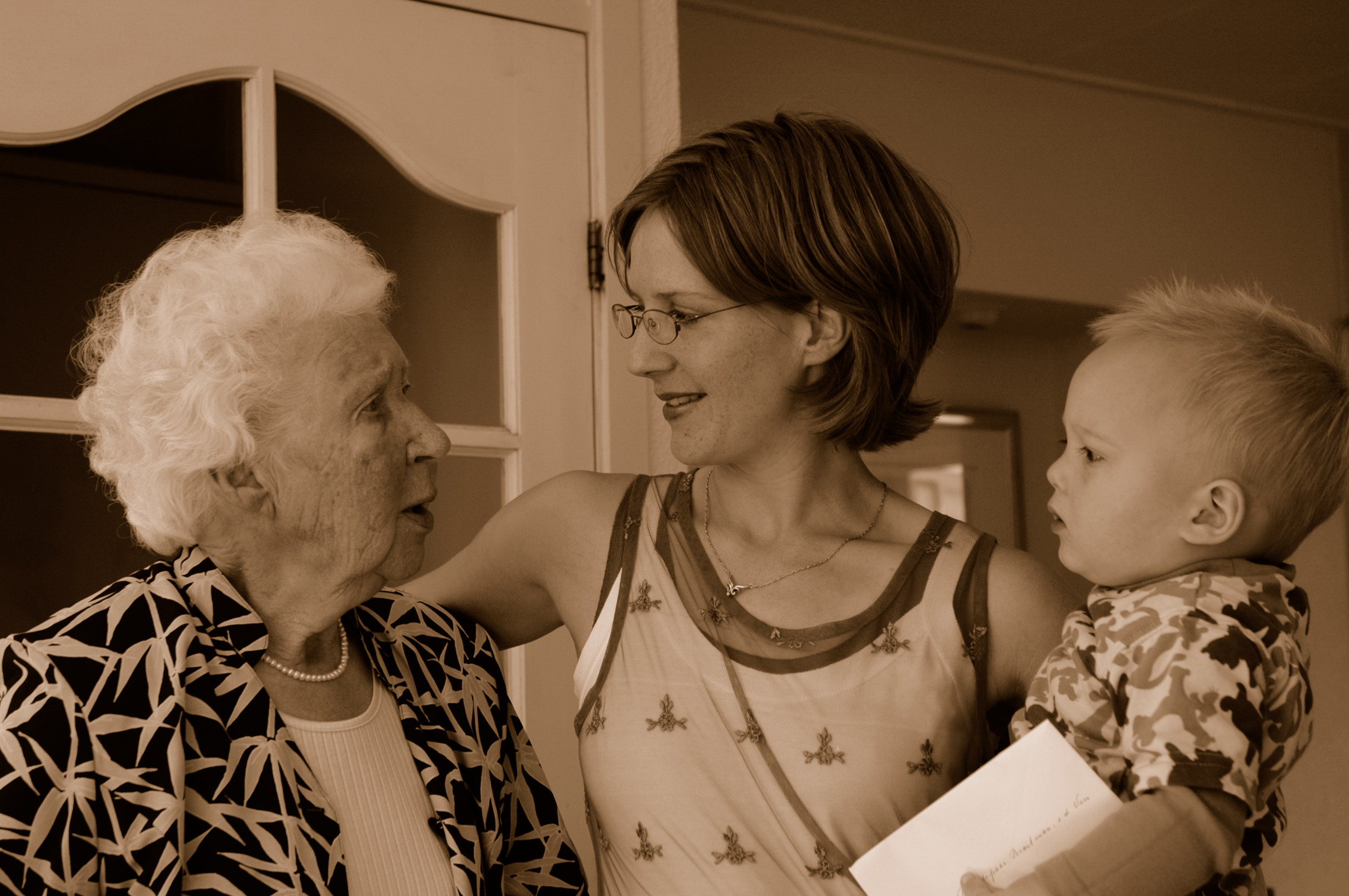 A woman holding a baby smiles while talking to an elderly woman indoors.