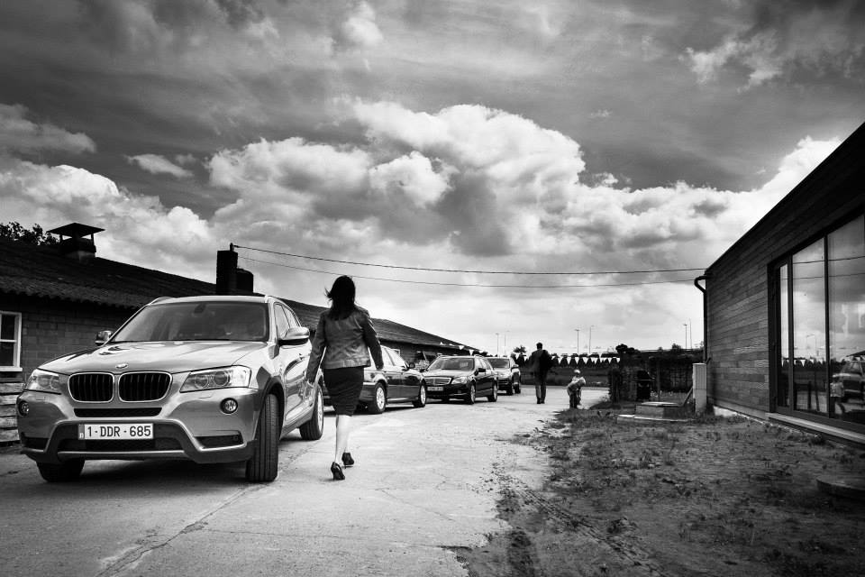 A woman in a dress and jacket walks toward a group of people near parked cars at a wedding venue.