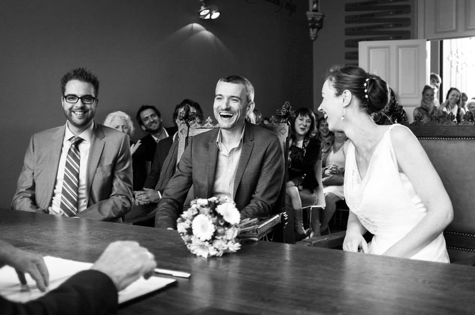 The bride and groom laugh joyfully during their wedding ceremony, surrounded by smiling guests in a lively atmosphere.