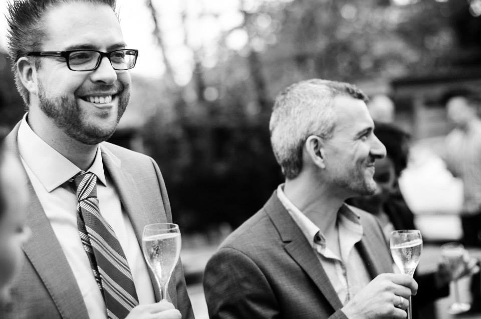 Two men in suits smile while holding champagne glasses at an outdoor wedding reception.