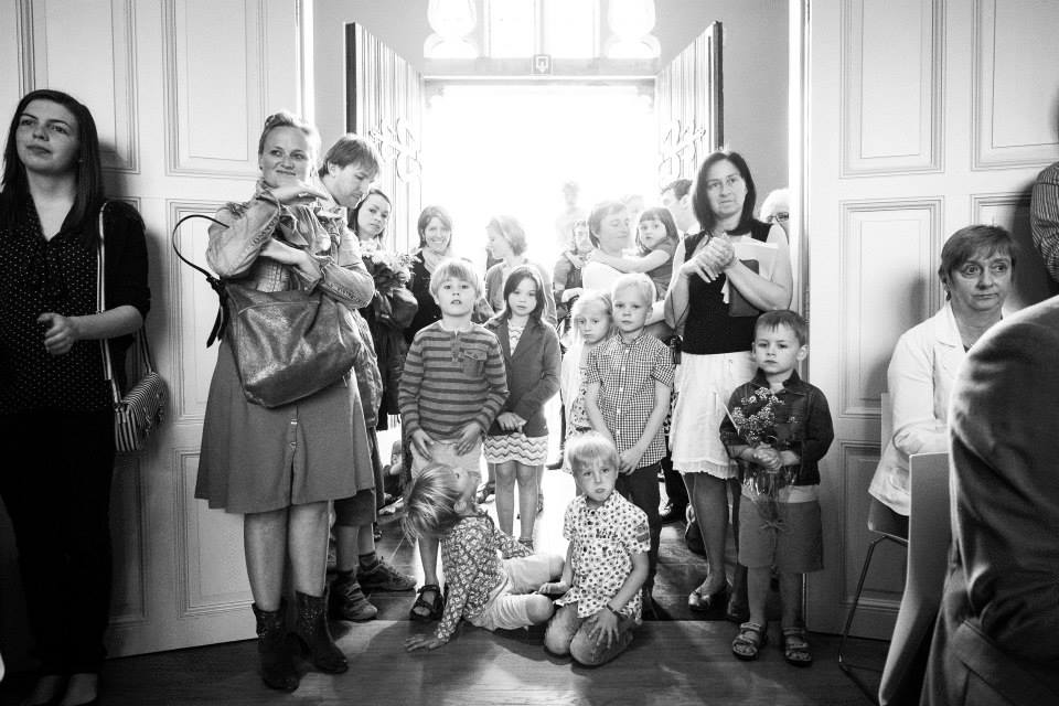 A group of wedding guests, including children, stand near an open door, watching and waiting with anticipation.