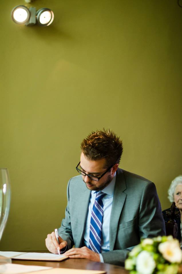 A man in a suit signs a document at a wedding ceremony, with an older guest seated in the background.