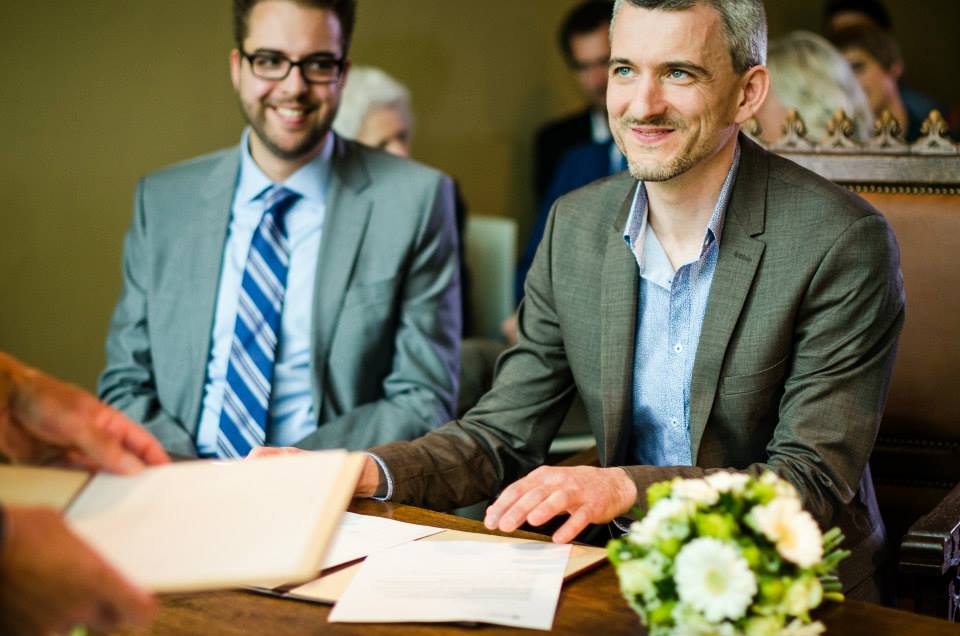 A smiling groom in a suit sits at a table, signing a document, with another man beside him.