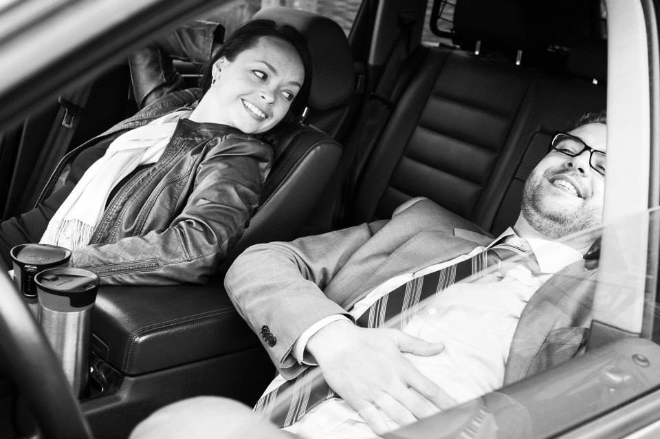 A man in a suit reclines in a car seat, smiling, while a woman beside him looks at him.