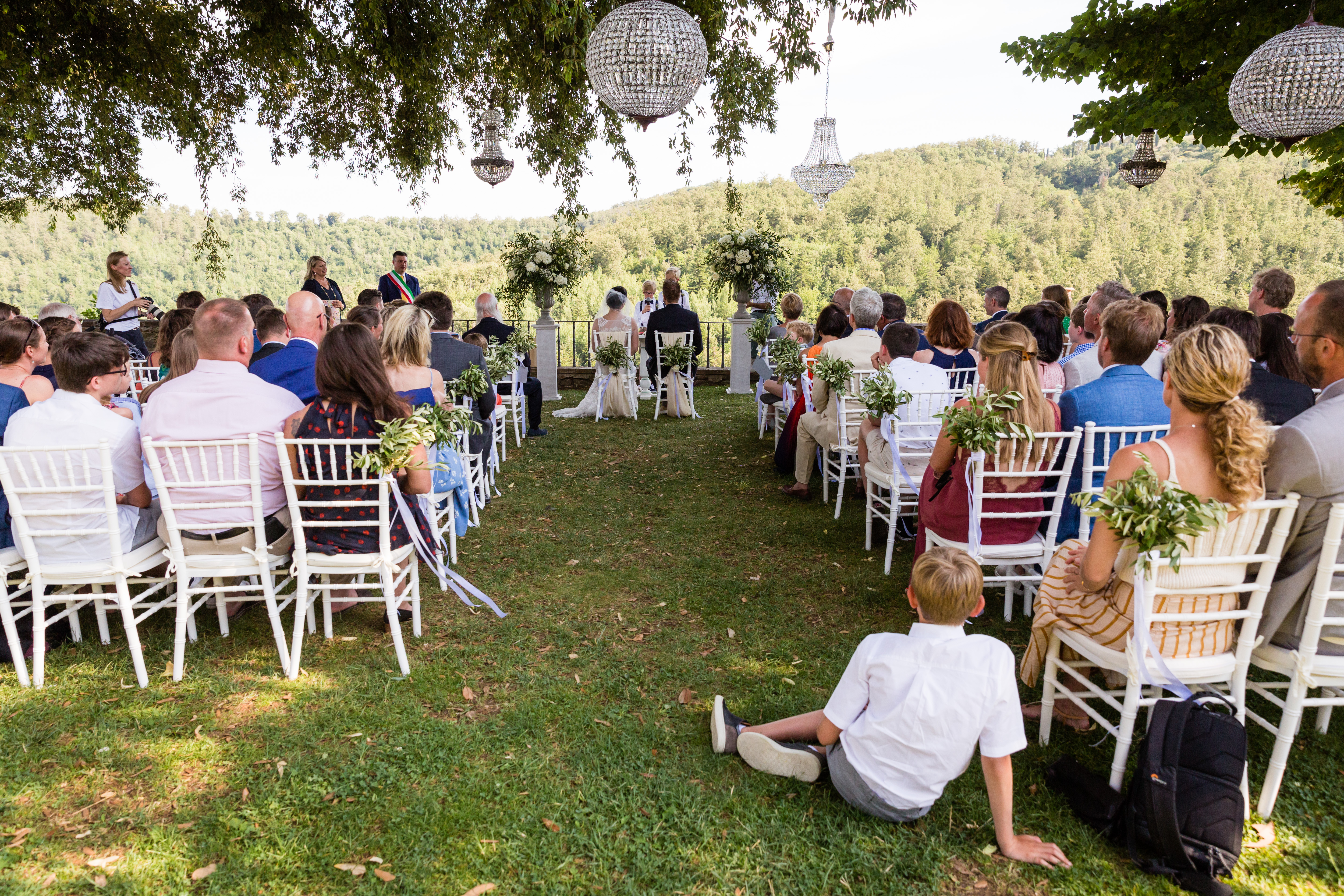 Wedding ceremony taking place outdoors with guests seated, facing the couple and officiant at the front.