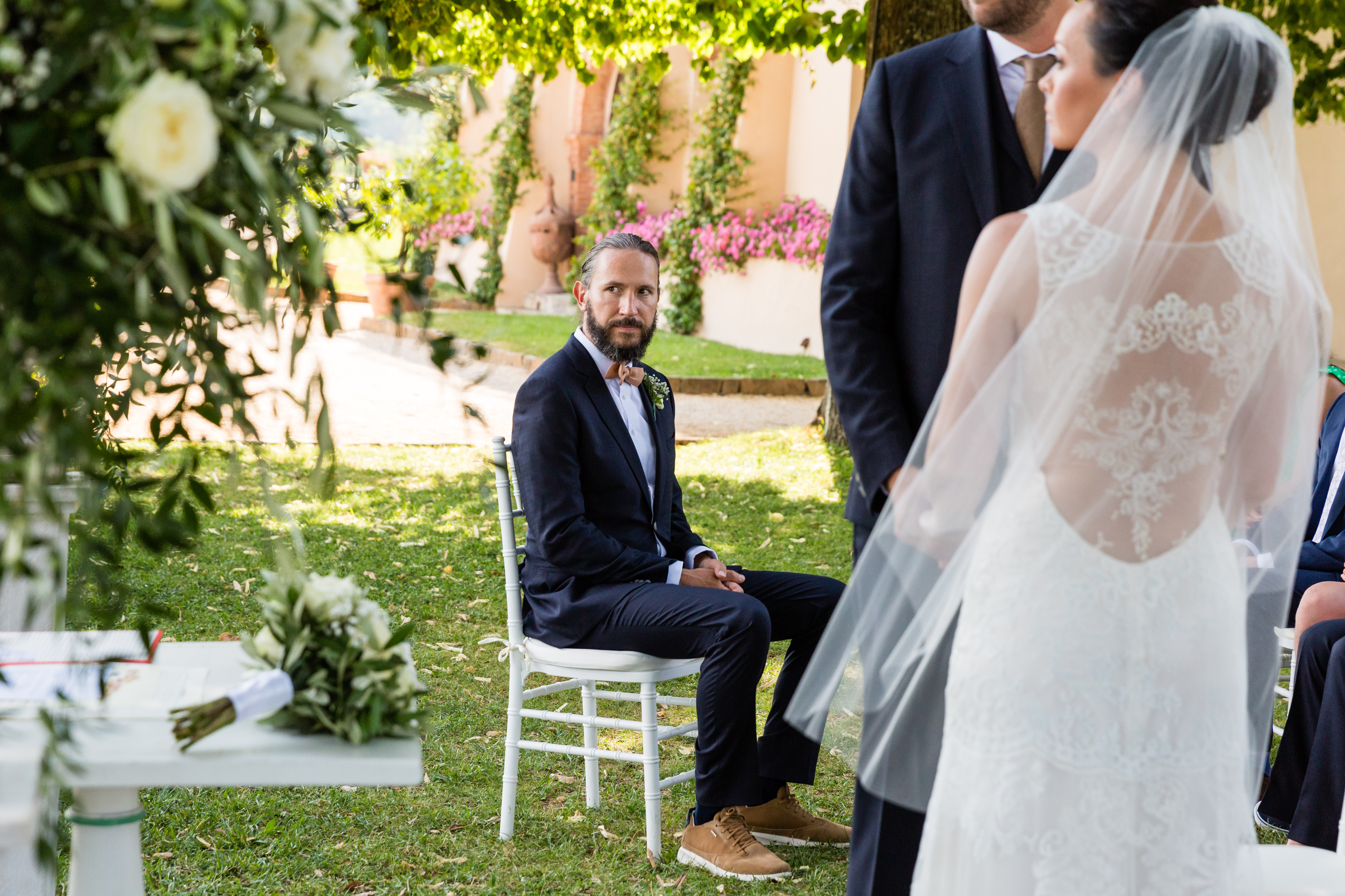 The groom sits on a chair, looking towards the bride, who stands in a white wedding dress with a veil.