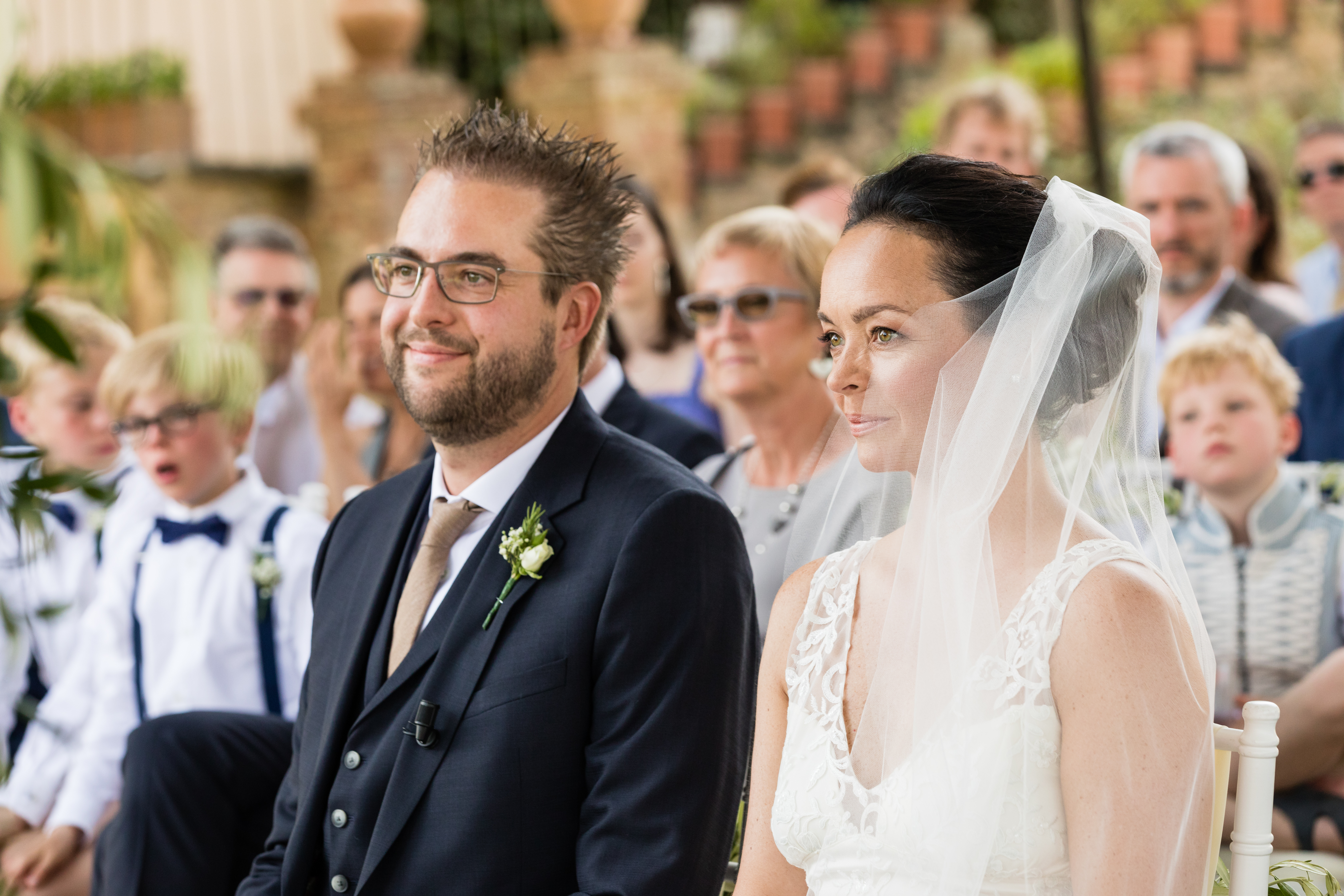 The bride and groom sit side by side during their wedding ceremony, surrounded by guests.