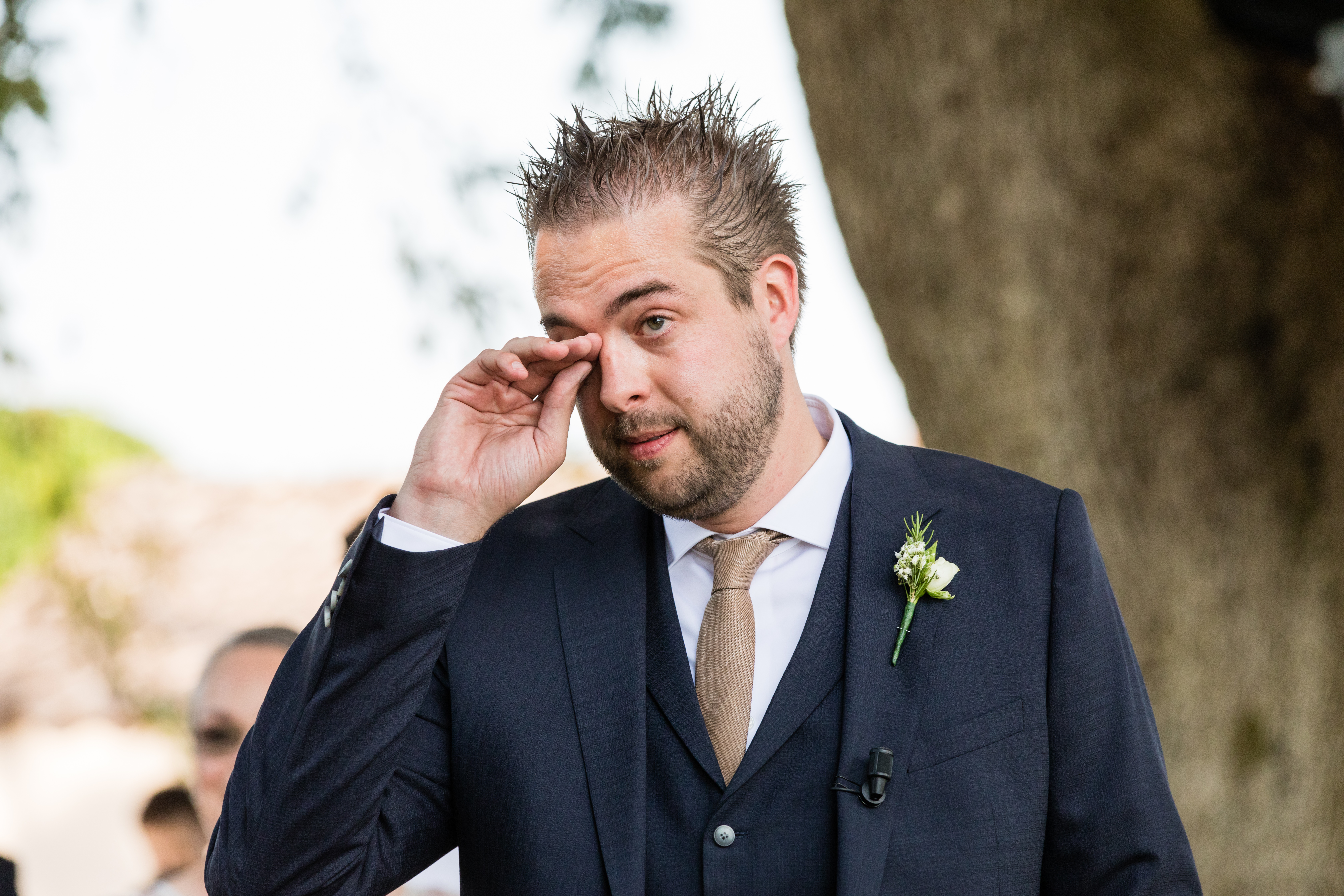 A groom in a dark suit wipes his eye during the wedding ceremony, showing emotion.