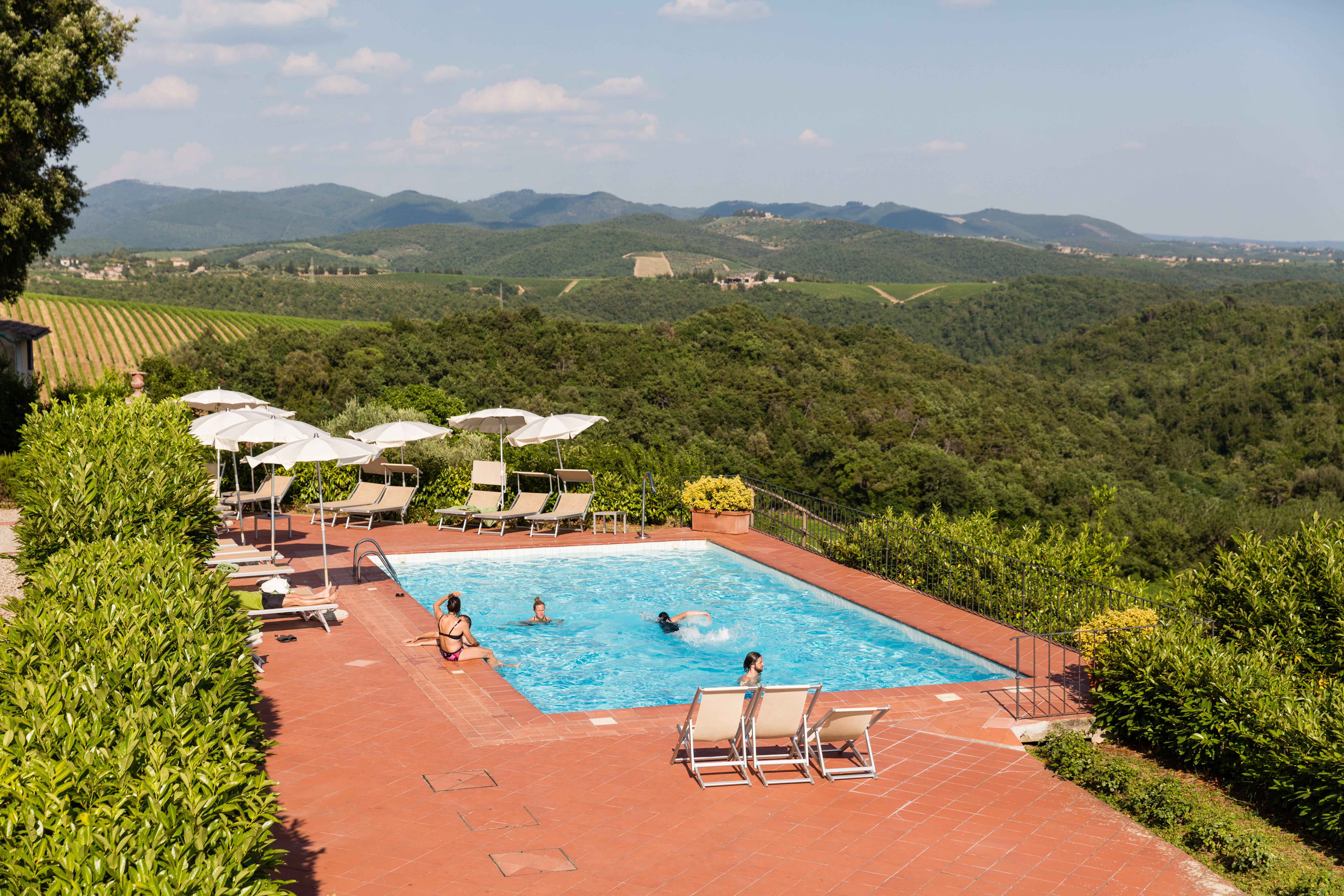 A few people relax in and around a swimming pool with a scenic hillside view in the background.
