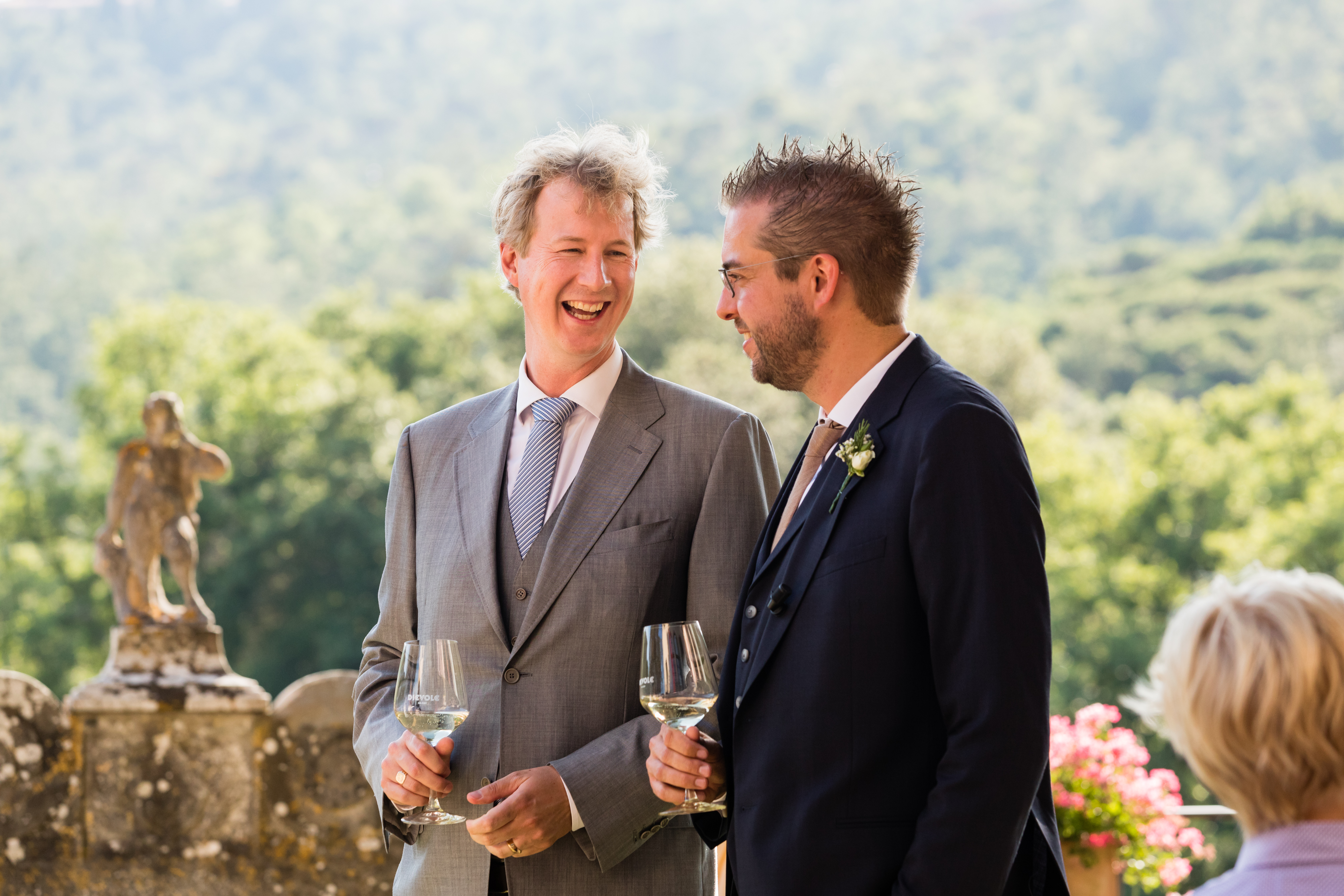 Two men in suits stand outside, holding wine glasses and smiling while engaged in conversation at a wedding.