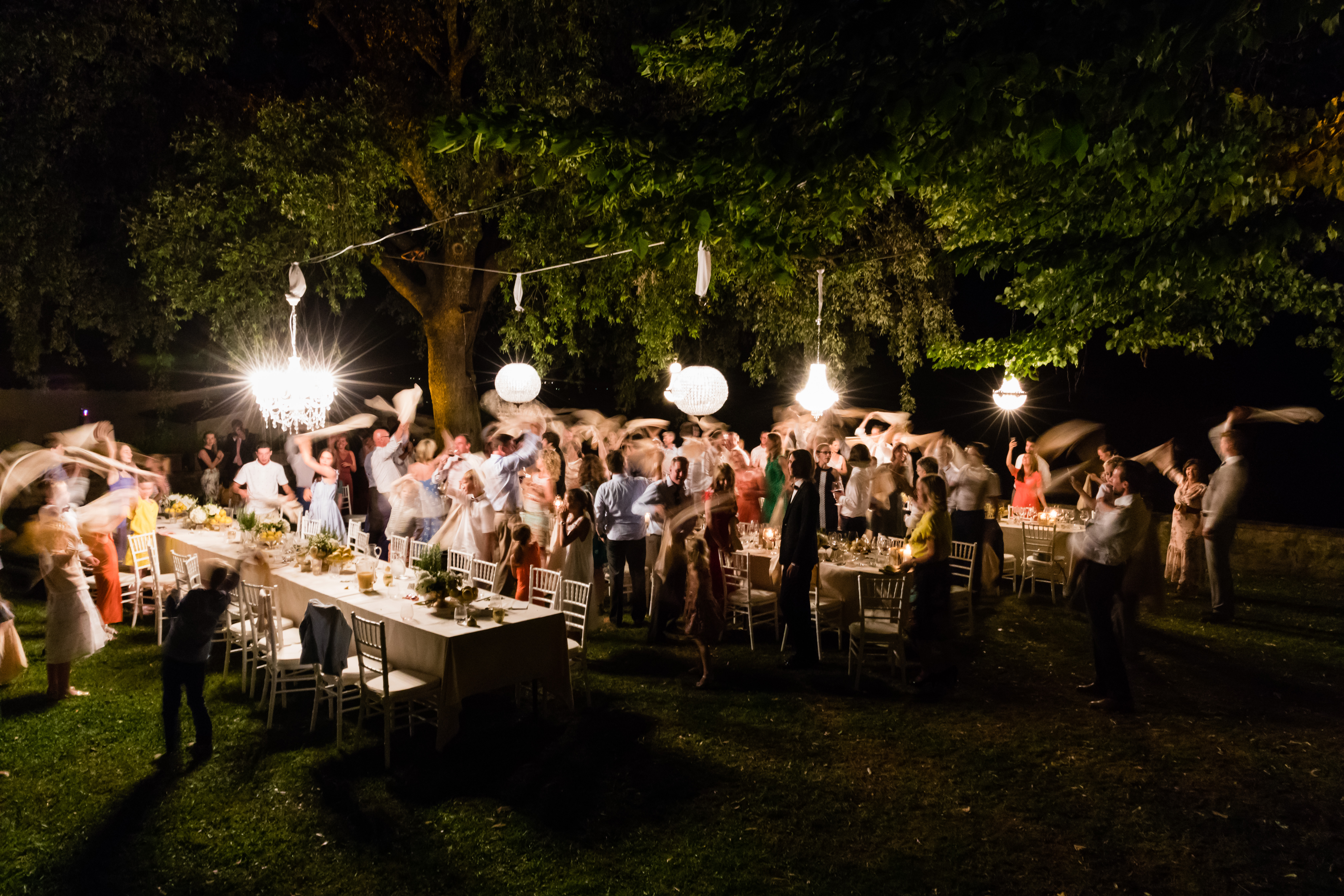 Wedding guests stand and wave napkins in the air during an outdoor dinner celebration under hanging lights.