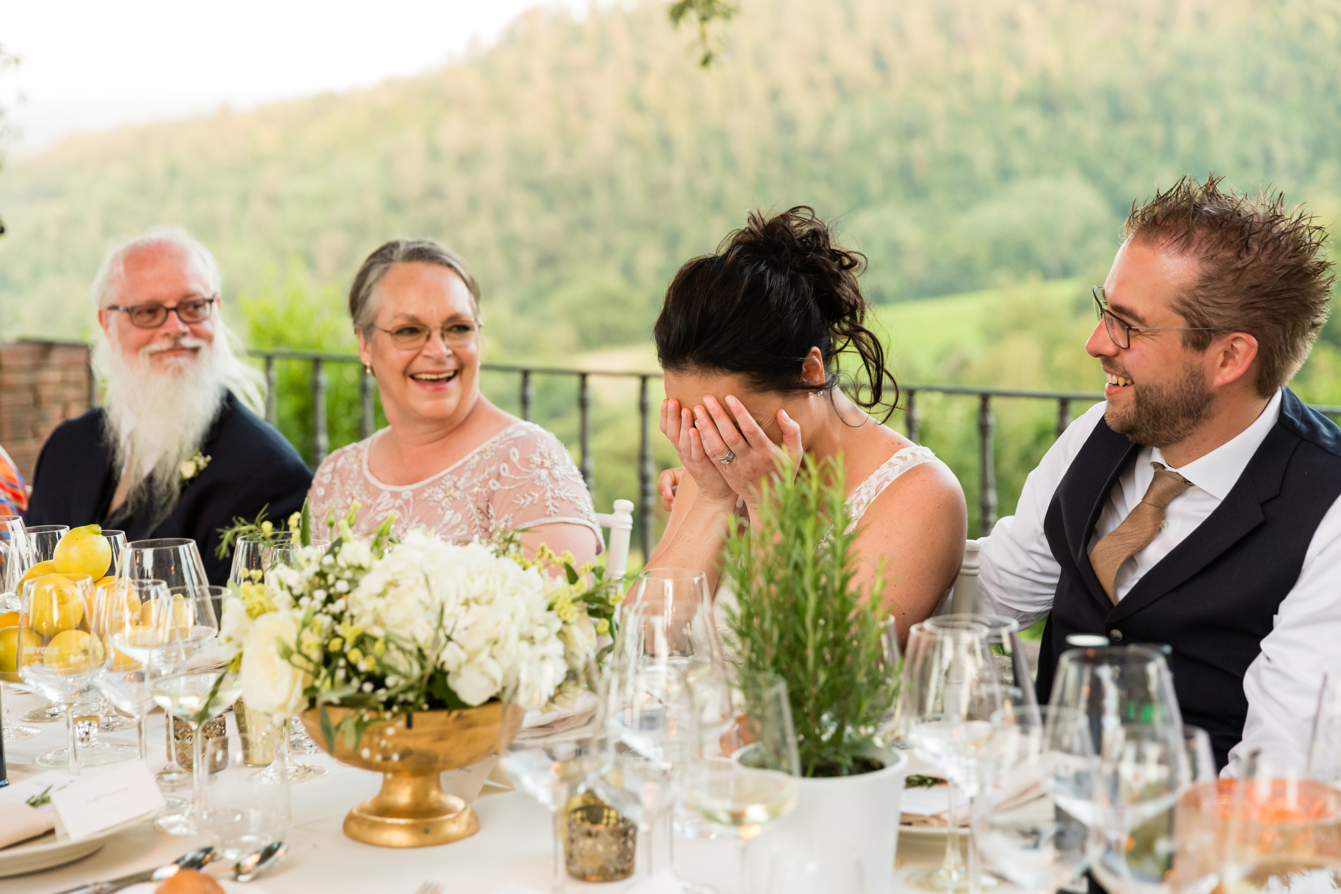 The bride covers her face while laughing at the dinner table, surrounded by smiling guests.