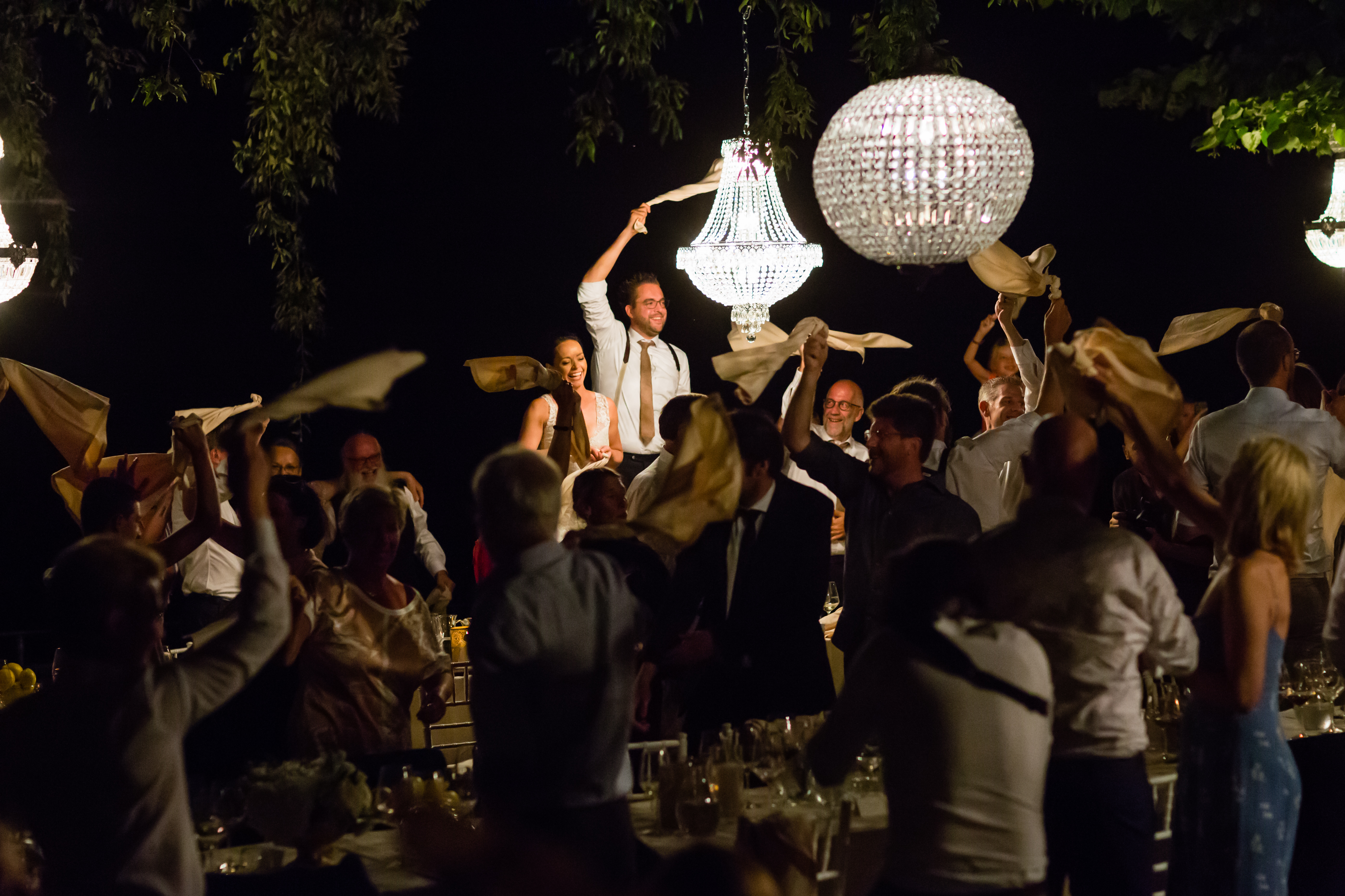 Wedding guests wave napkins in the air as the bride and groom stand and celebrate during dinner.