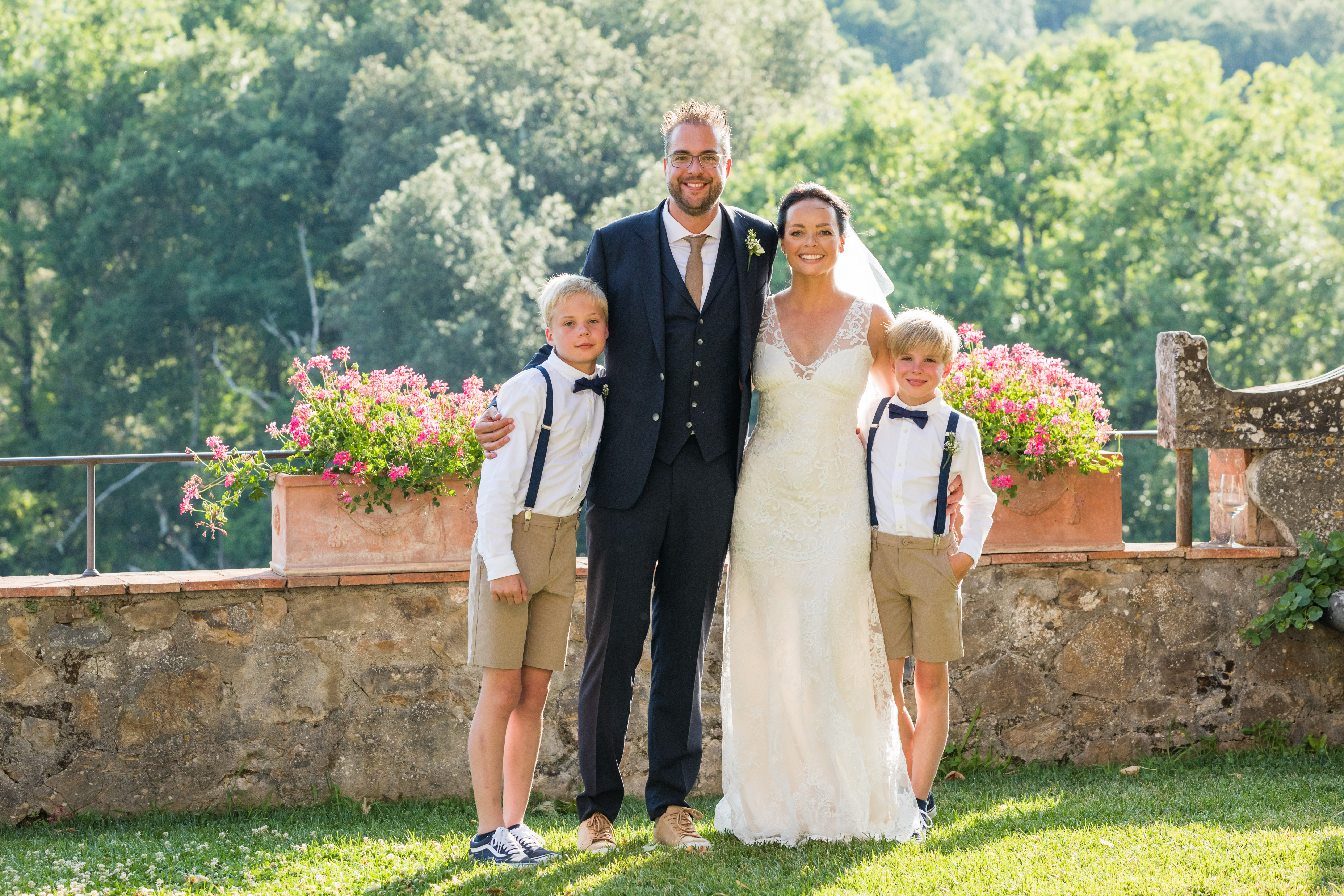 Bride and groom stand smiling with two young boys in suspenders, posing together for a wedding photo.