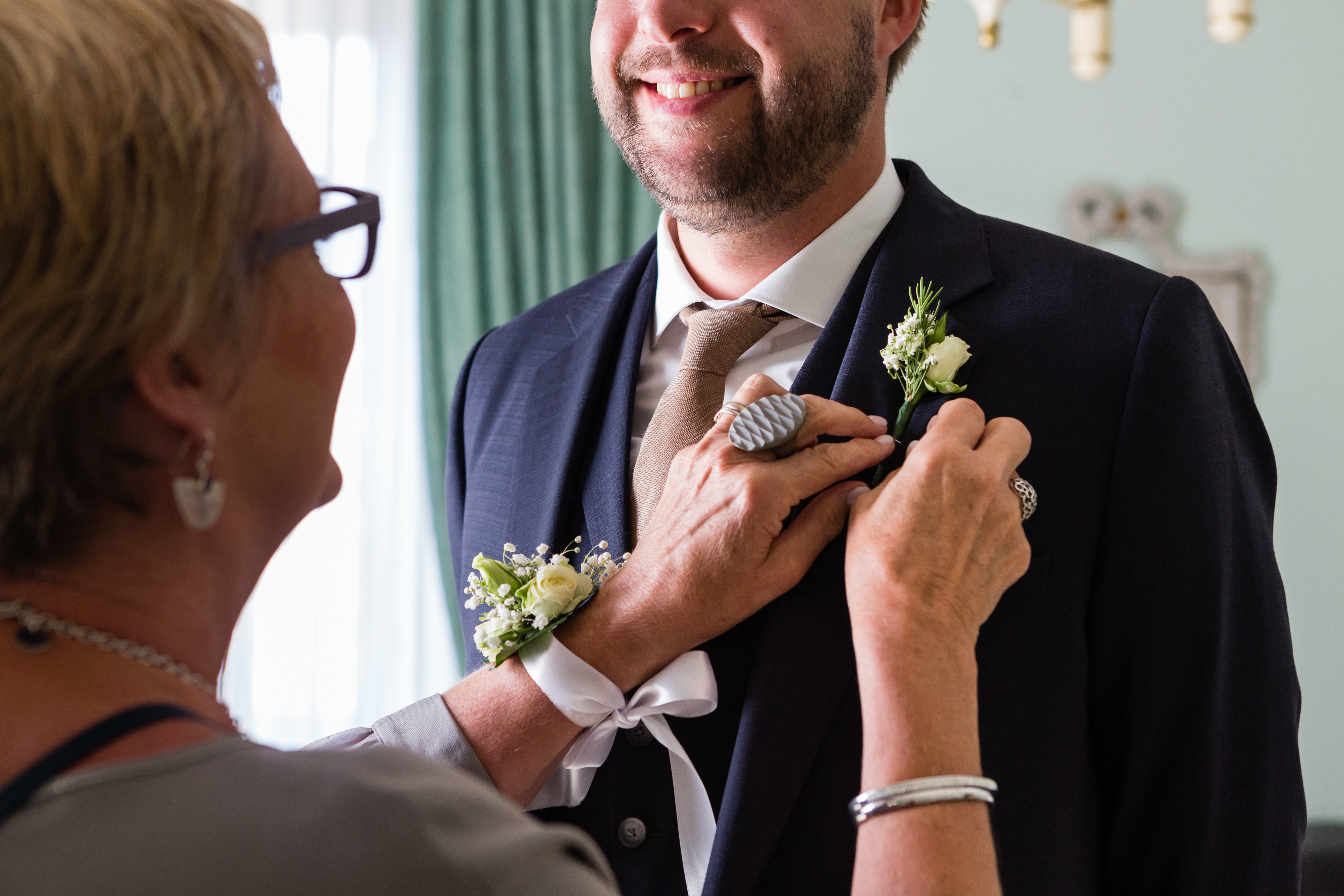 A woman pins a boutonniere on a smiling groom's suit jacket as they prepare for the wedding.