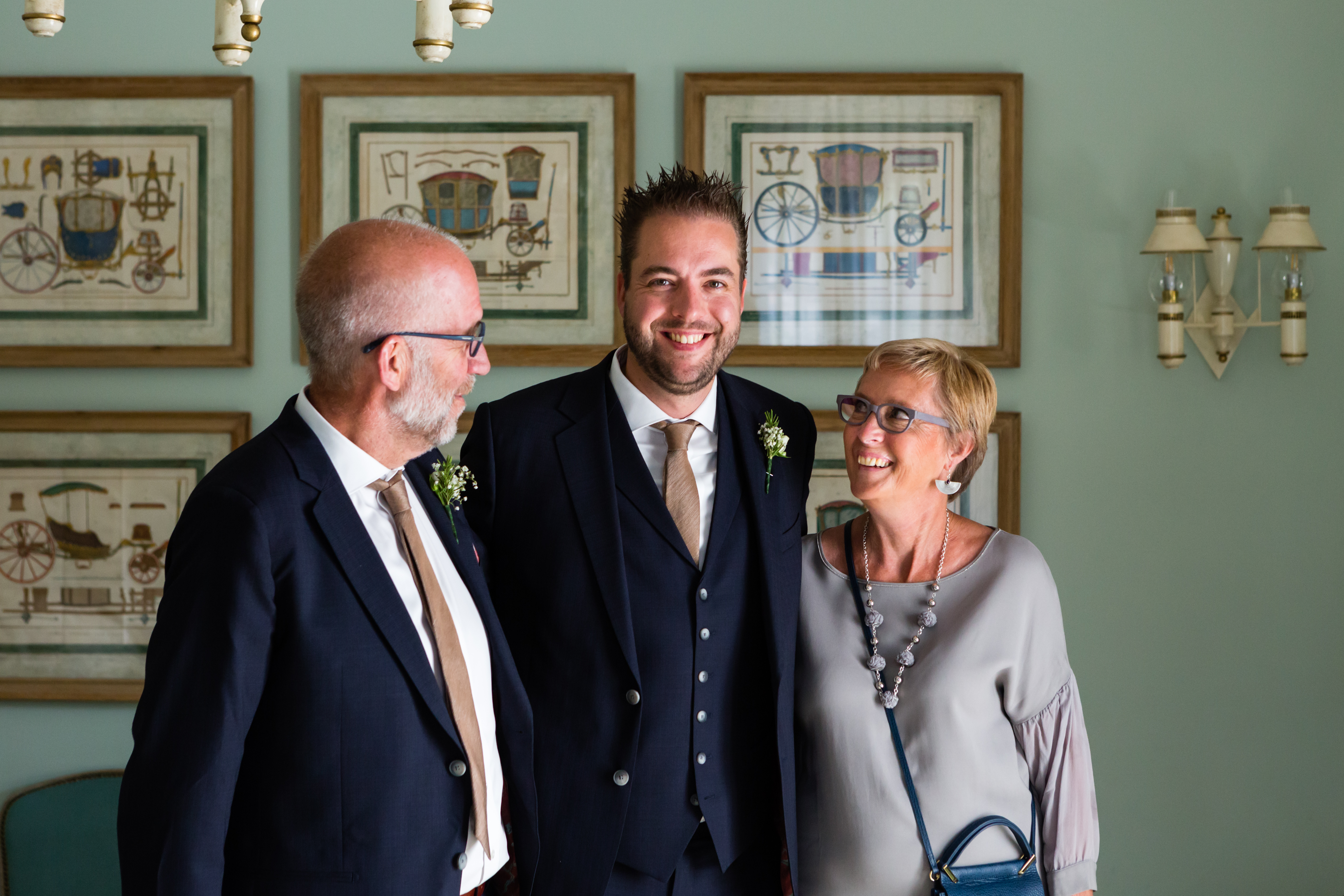 A groom in a suit smiles while standing between two older adults, likely family, before the wedding.