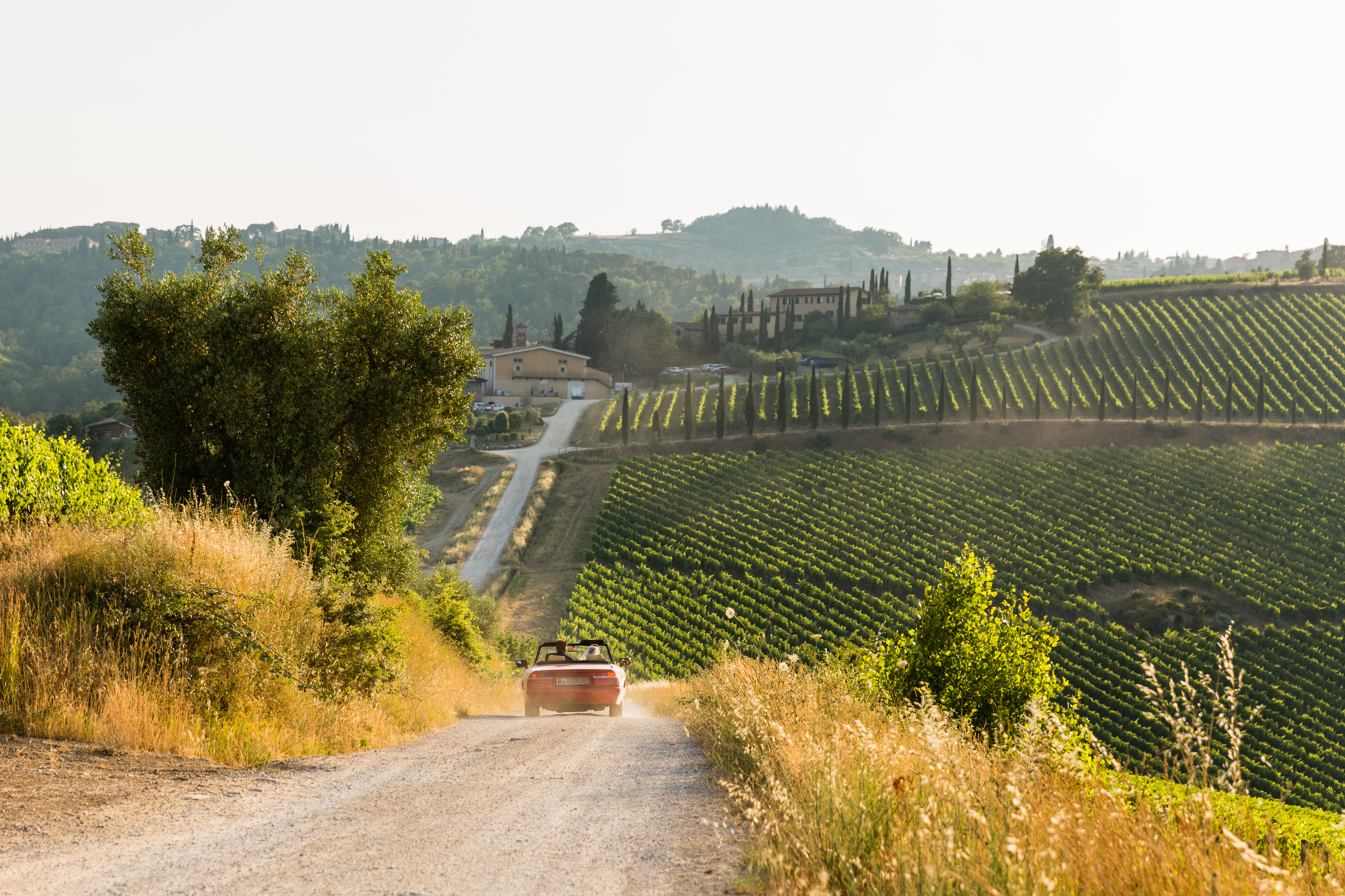 A couple drives a red convertible down a dirt road through a vineyard, leaving a trail of dust behind.