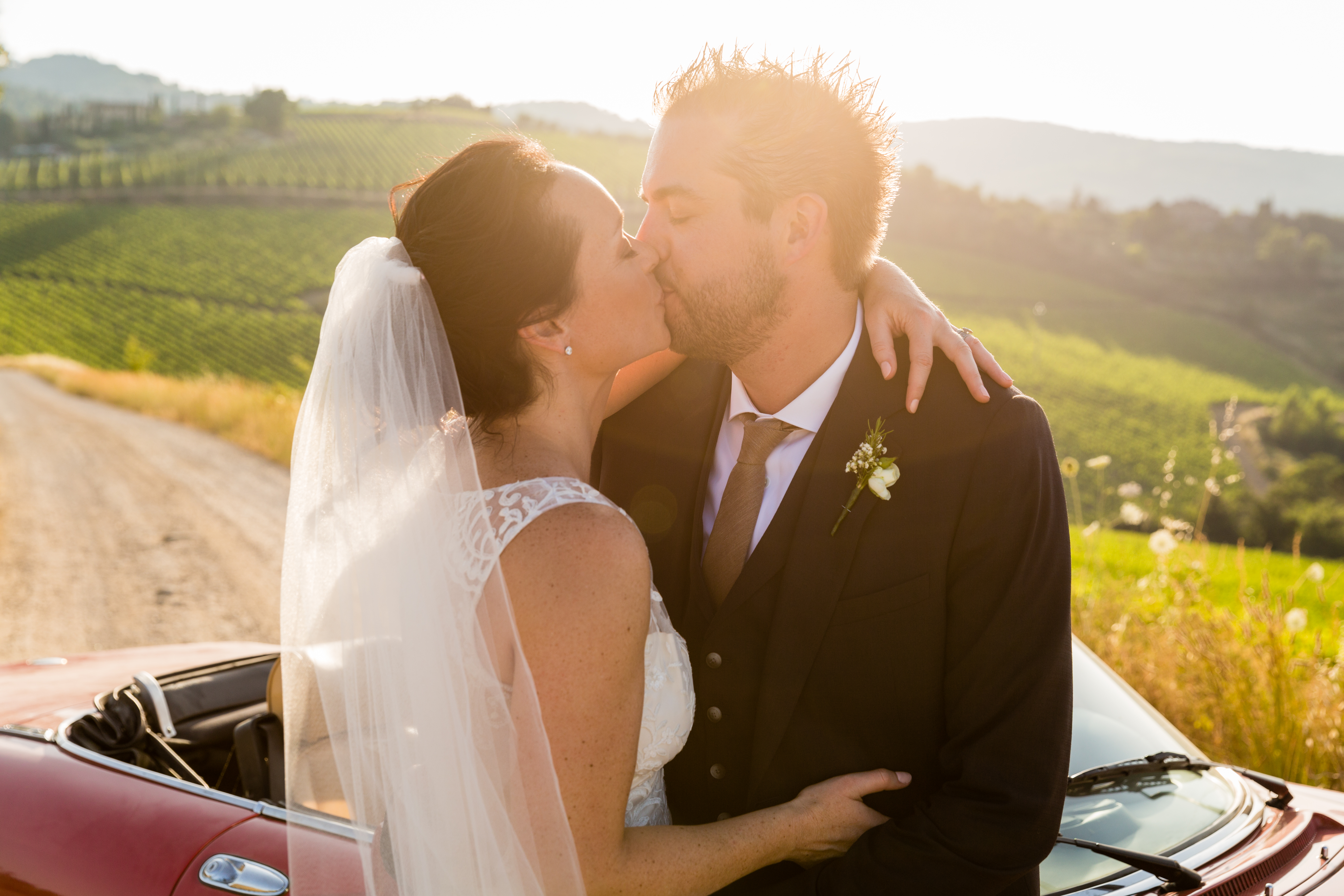 A bride and groom share a kiss in a vineyard, standing beside a red convertible car.