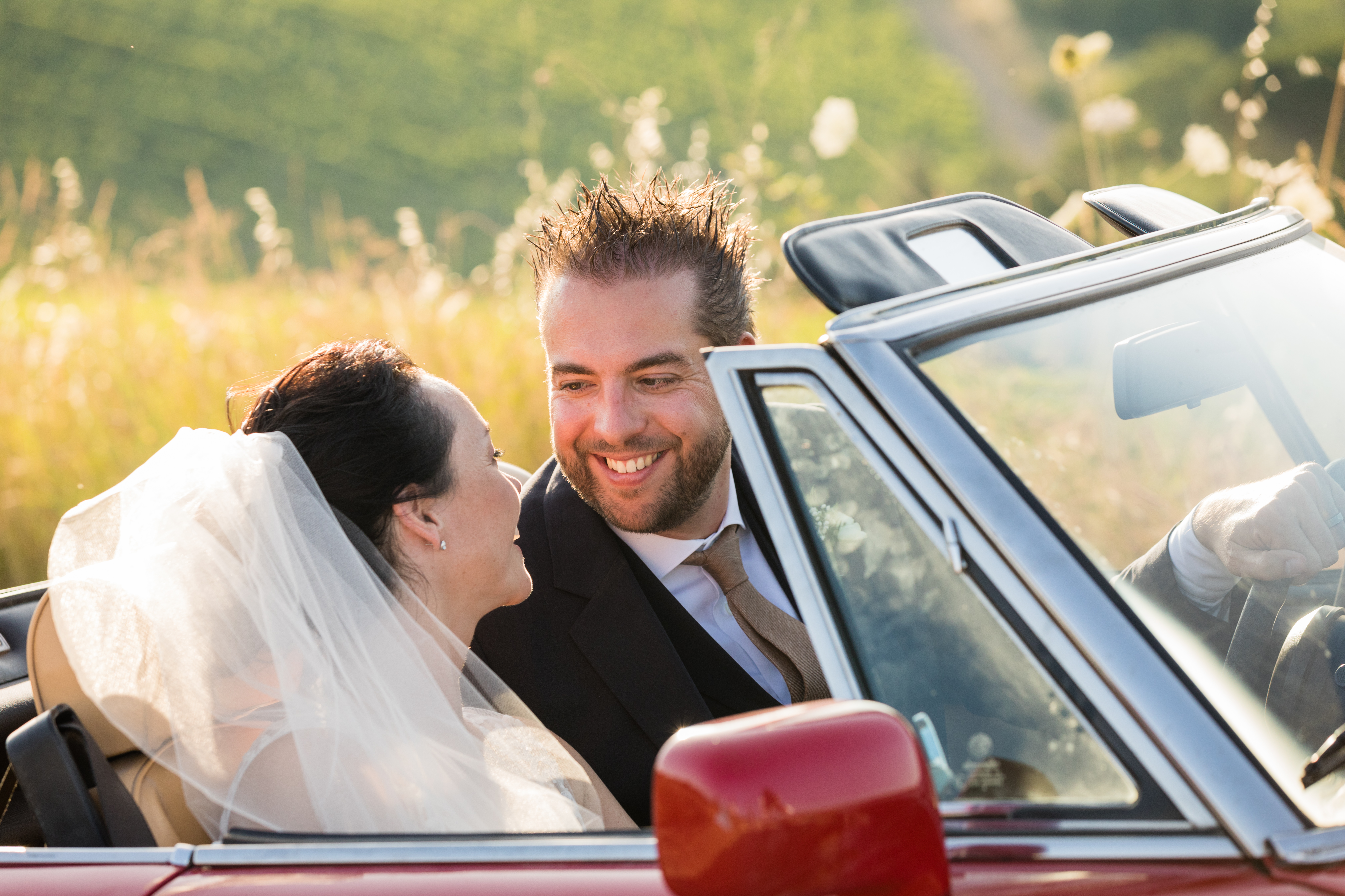 A bride and groom smile at each other while sitting in a red convertible with the top down.