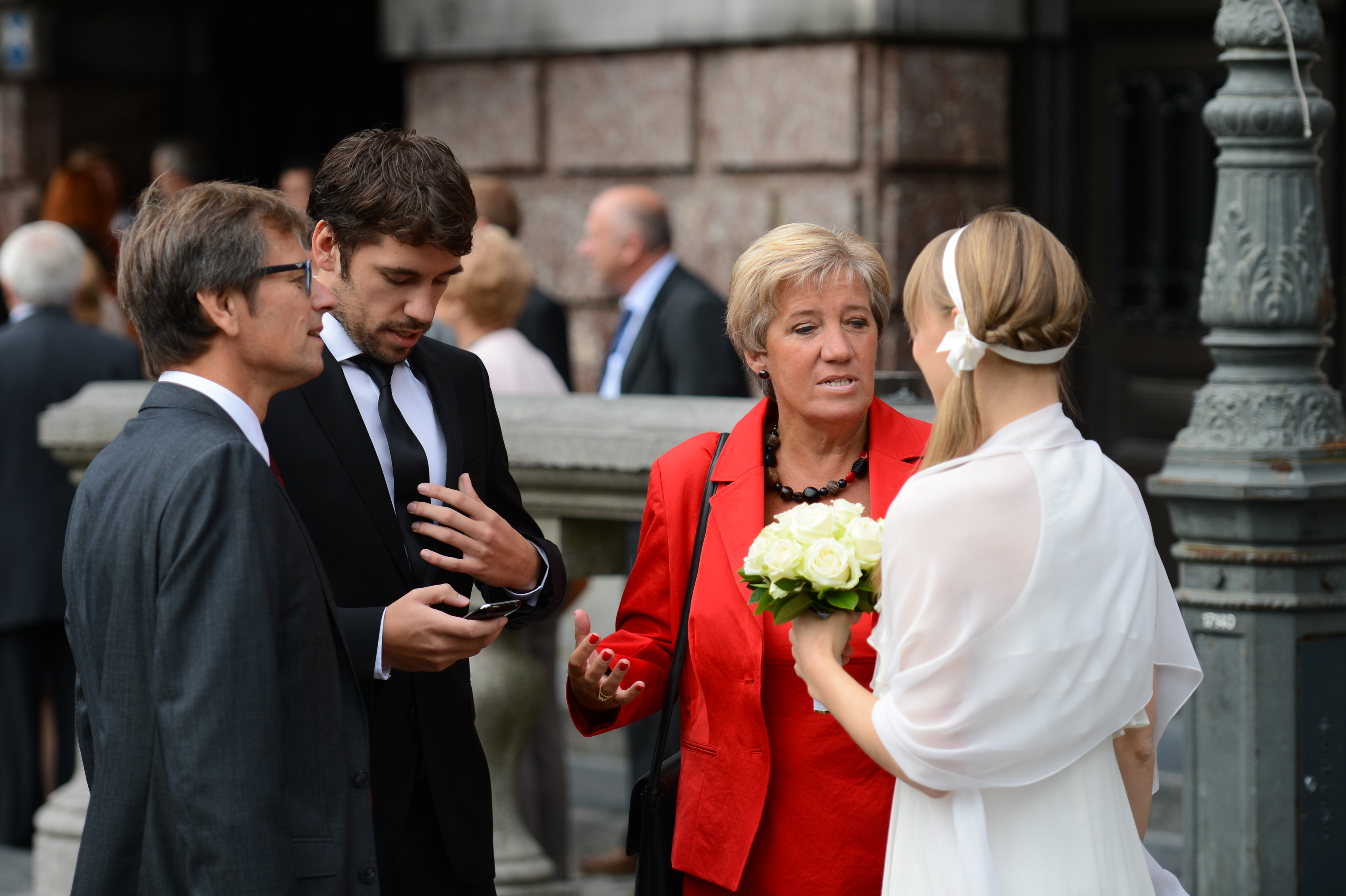 A bride holding flowers talks with three well-dressed guests outside a building during a wedding gathering.