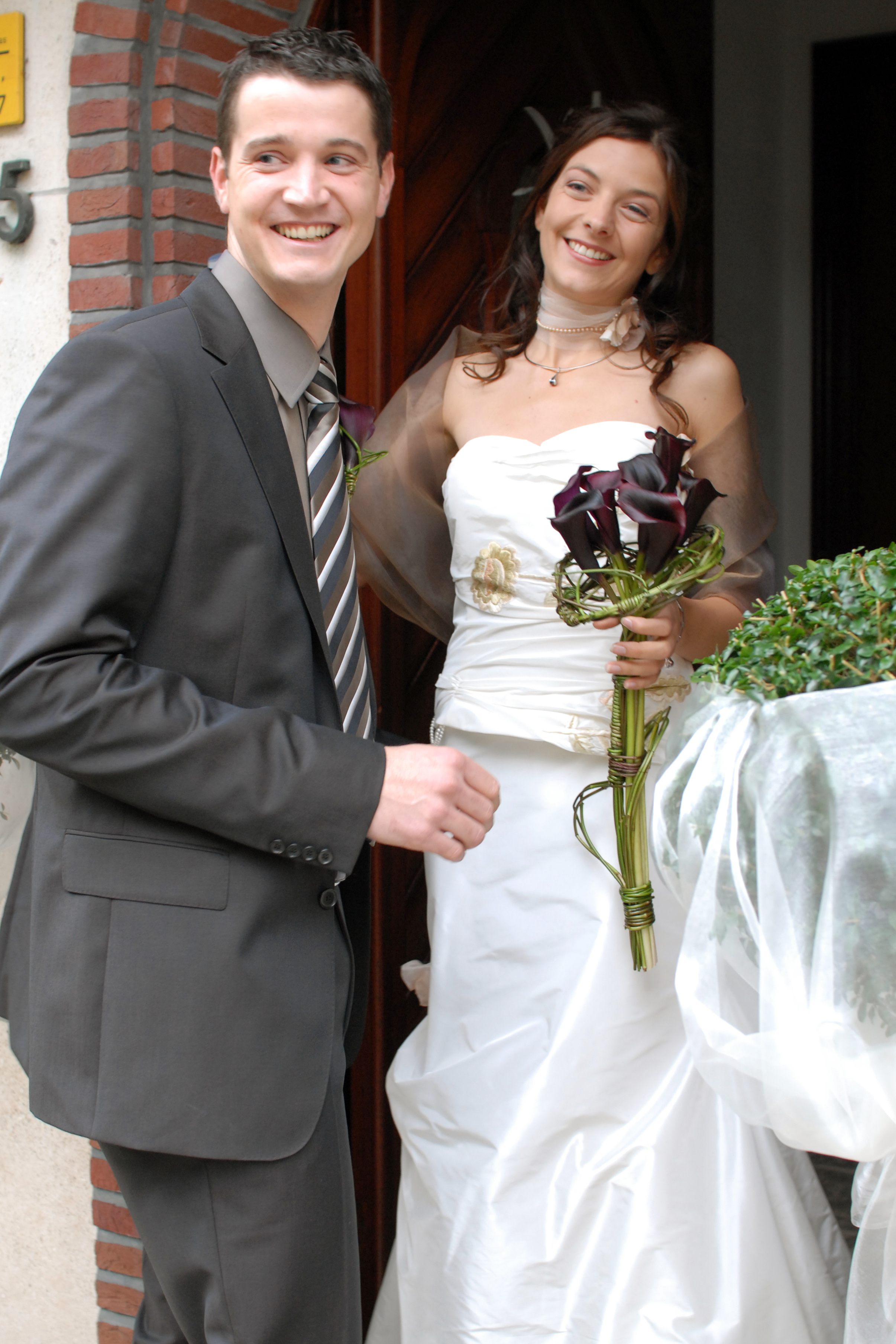 A smiling bride and groom stand together, with the bride holding a bouquet of dark flowers.