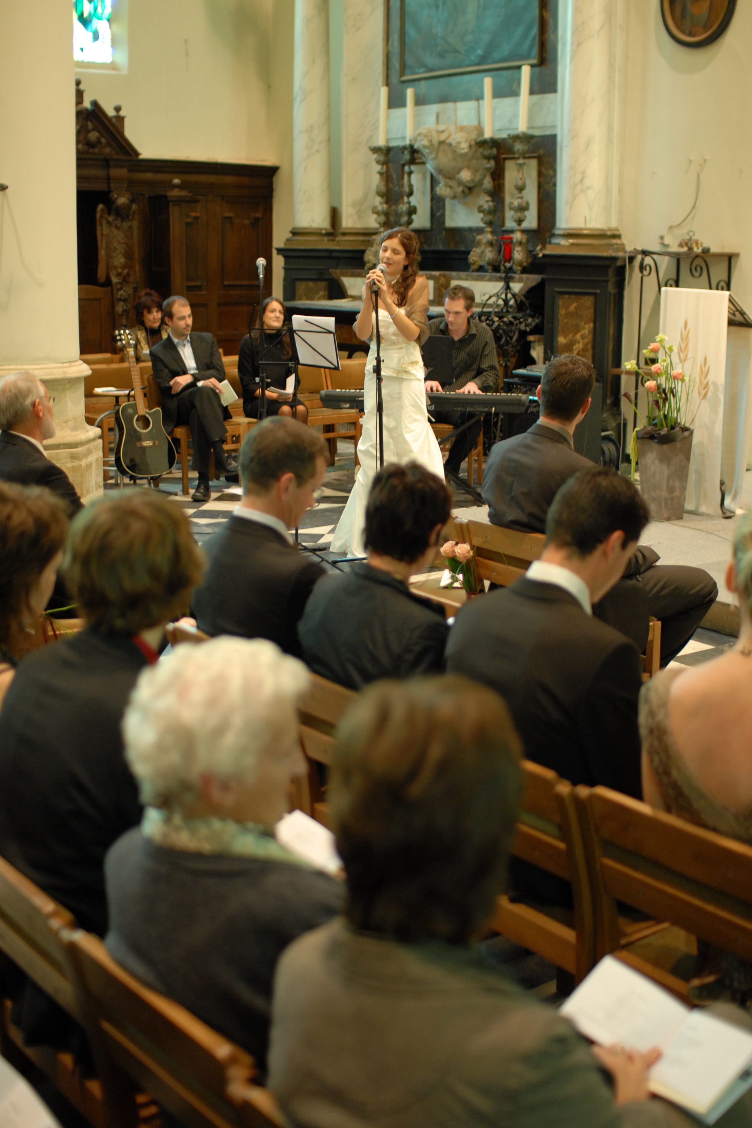 A woman in a white dress sings into a microphone at a wedding ceremony, with guests seated in pews.