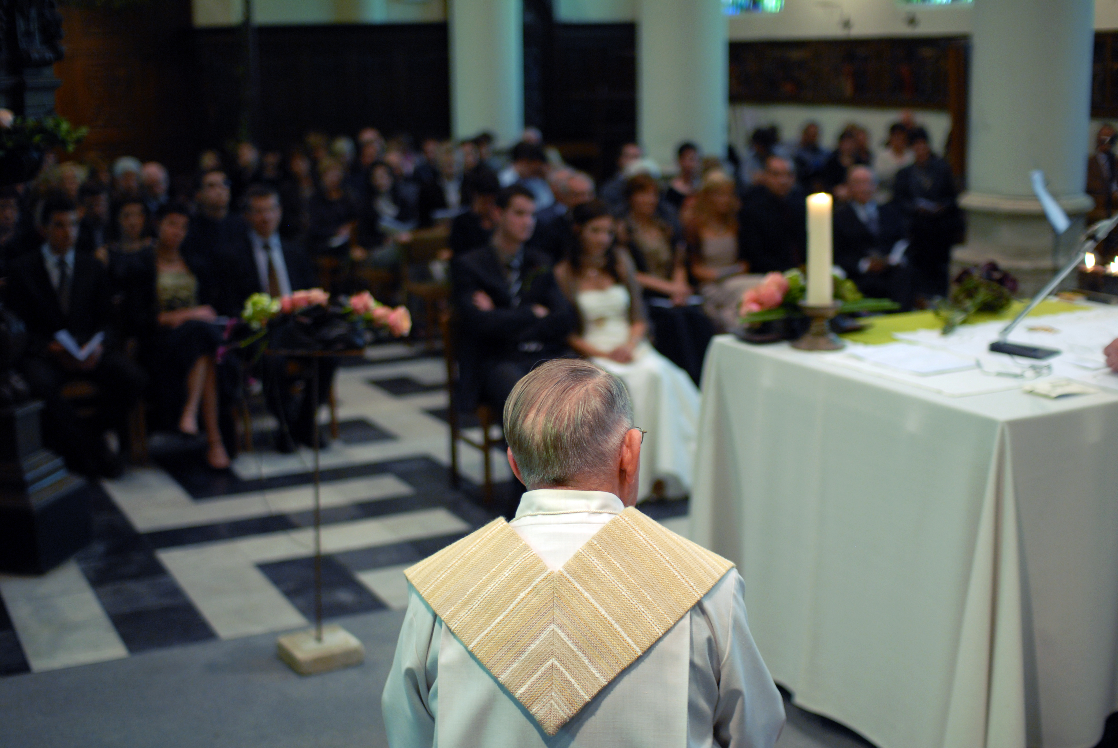 A priest stands at the altar during a wedding ceremony, with the bride and guests seated in the background.